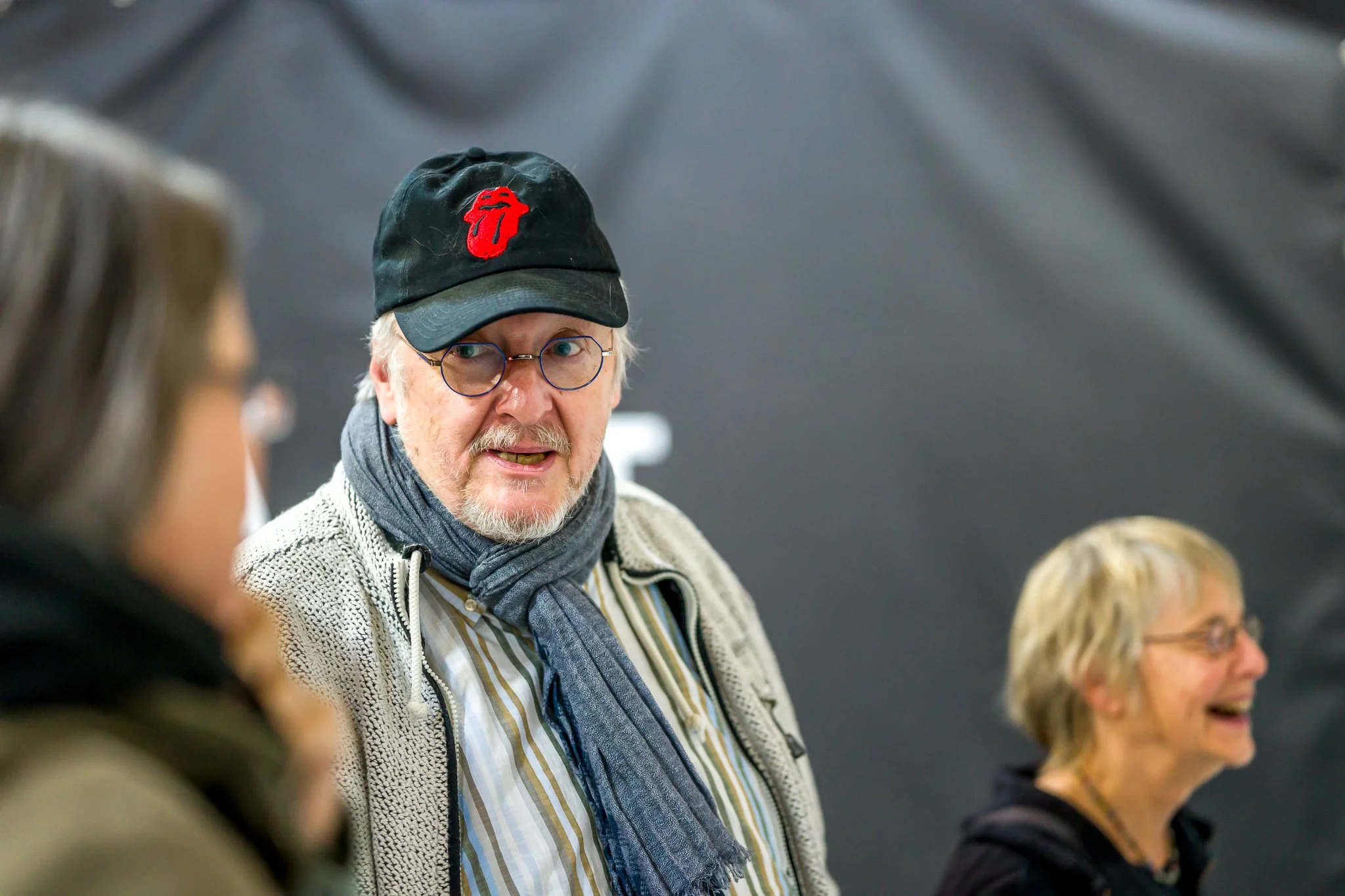 Un homme portant une casquette noire avec un logo rouge des Rolling Stones, des lunettes, une écharpe, discutant avec deux personnes, une autre femme souriante à l'arrière-plan.