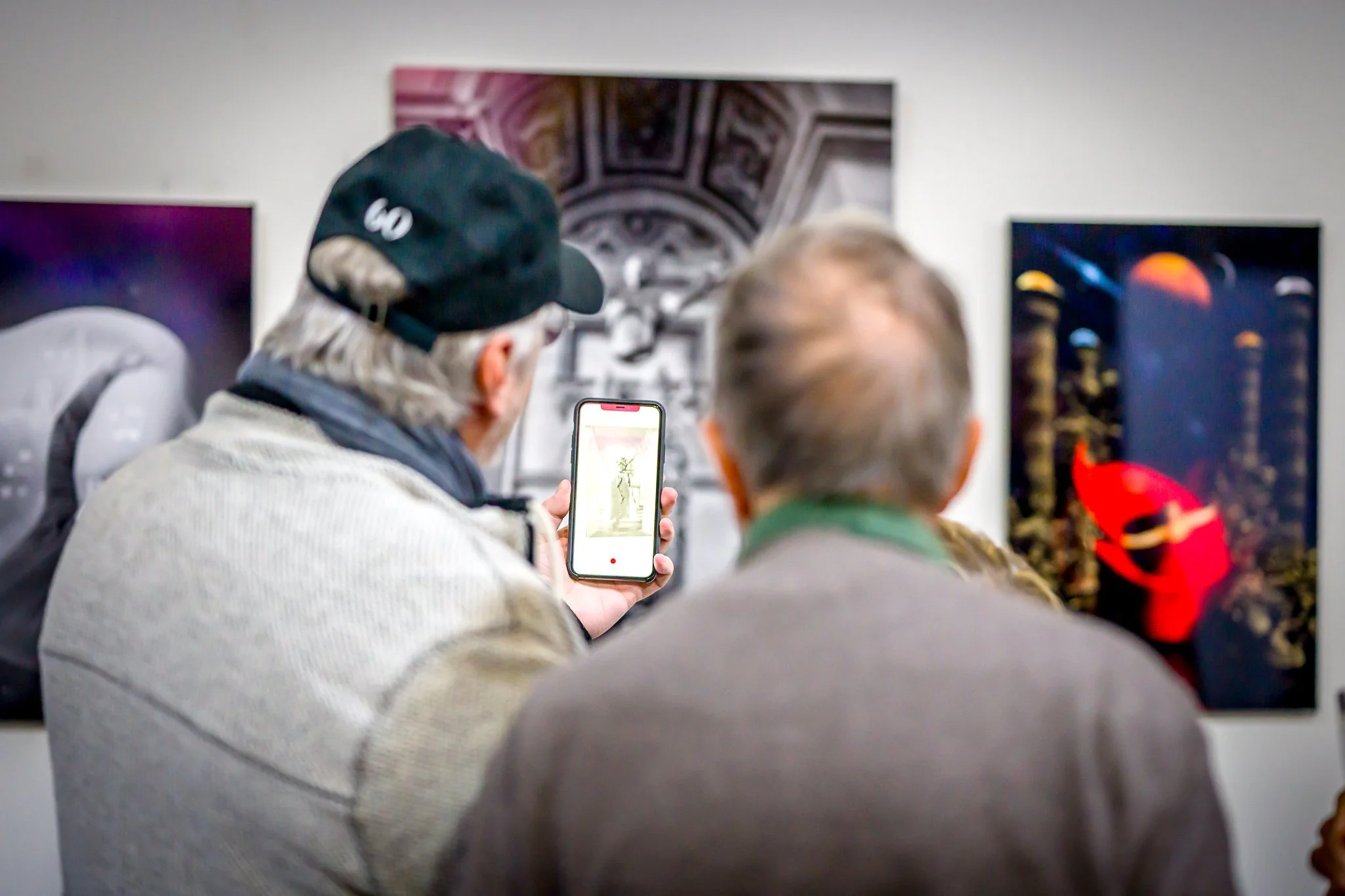 Deux hommes regardent des œuvres d'art sur un mur, l'un tient un smartphone montrant une image.
