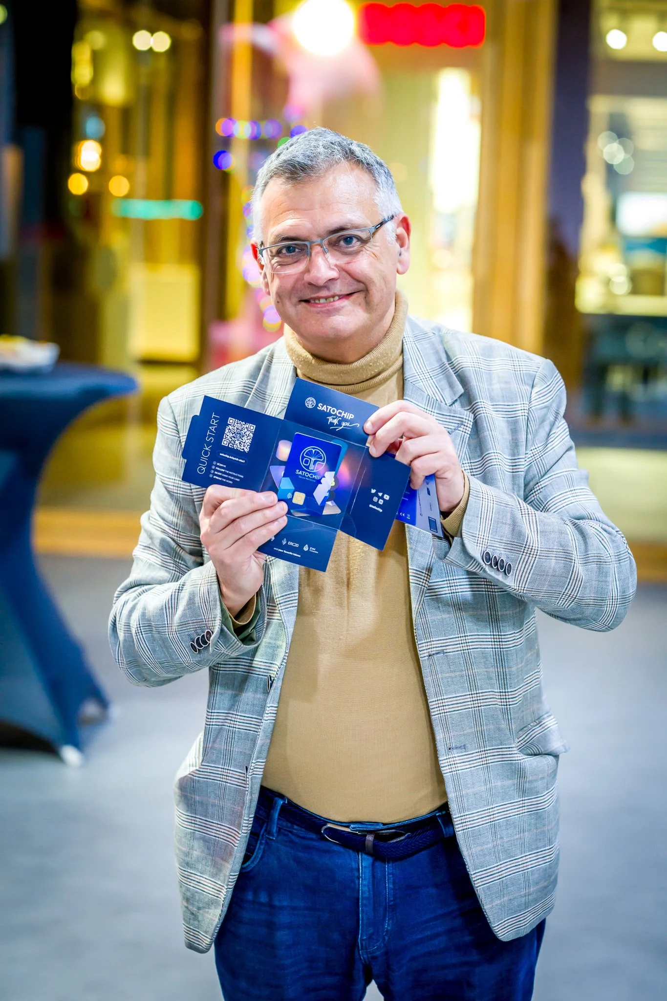 Un homme souriant tenant une brochure promotionnelle pour Satochip, portant une veste à carreaux et un pull beige, en intérieur avec des lumières floues en arrière-plan.