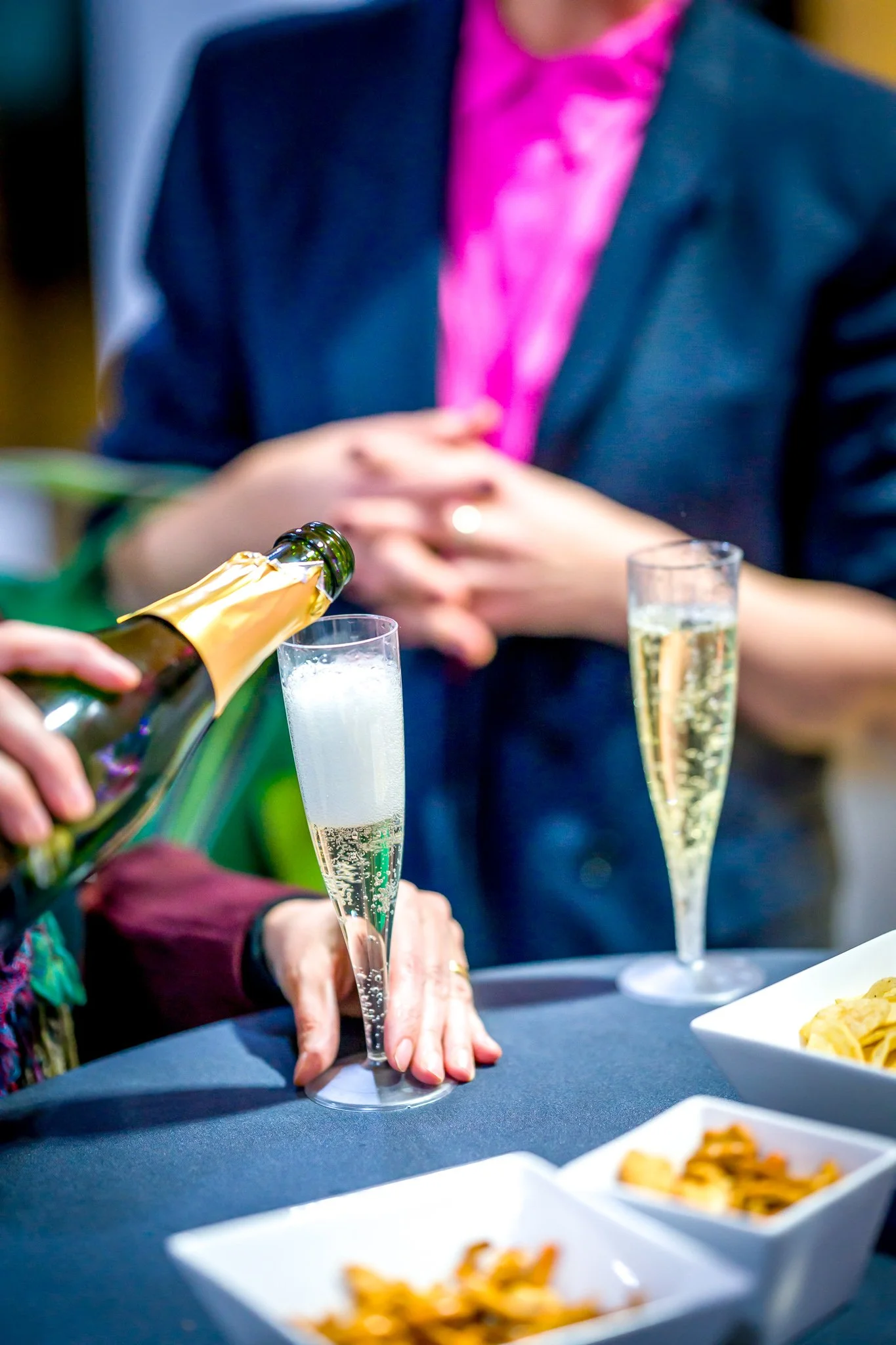 Une personne verse du champagne dans une flûte lors d'une réception ou célébration, avec des petits plats de snacks sur la table.