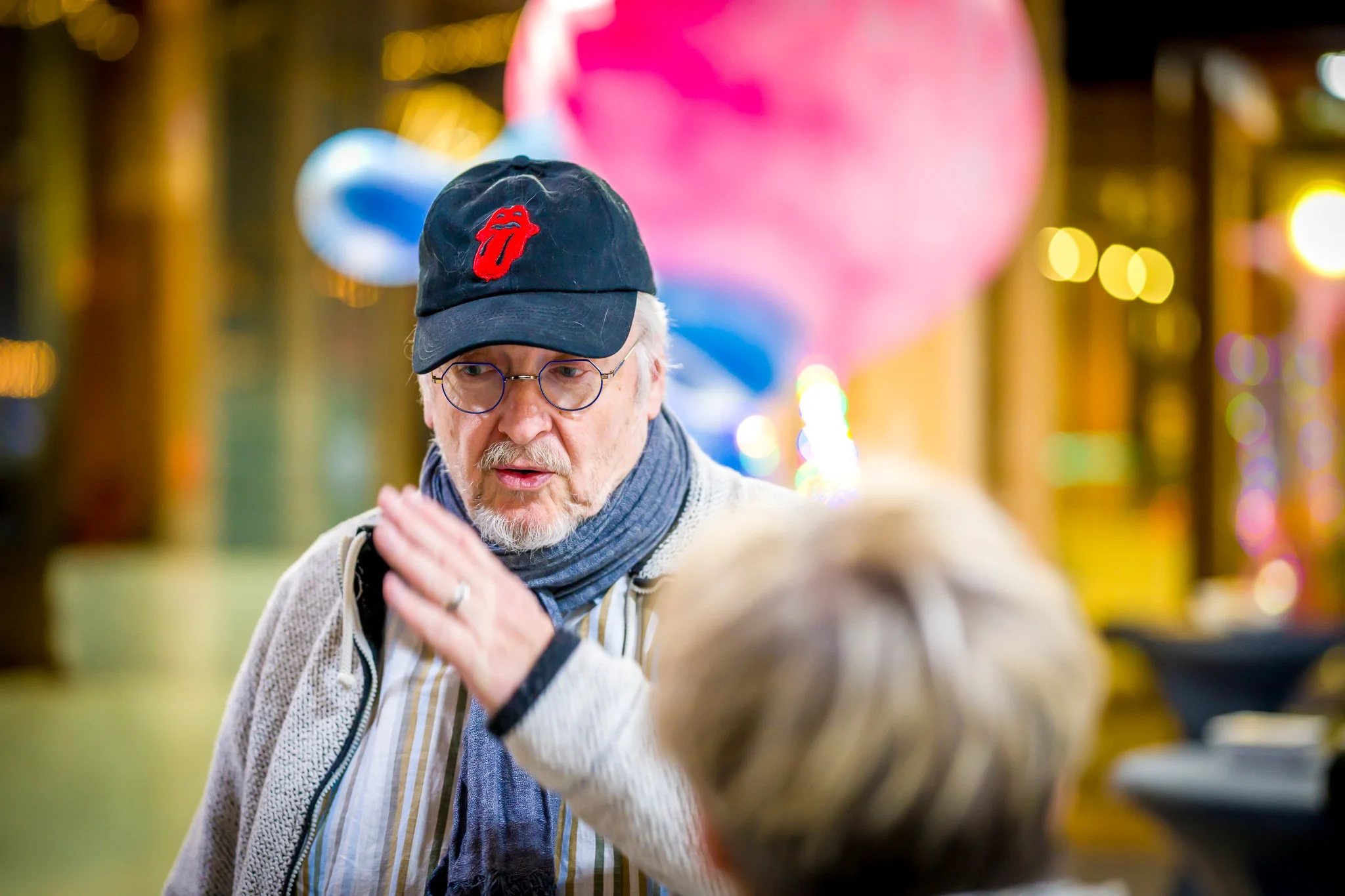 Deux personnes discutent dans un environnement intérieur coloré et flou. L'une des personnes porte une casquette noire avec un logo rouge, des lunettes et une écharpe grise. L’environnement est illuminé avec des lumières et des couleurs vives.