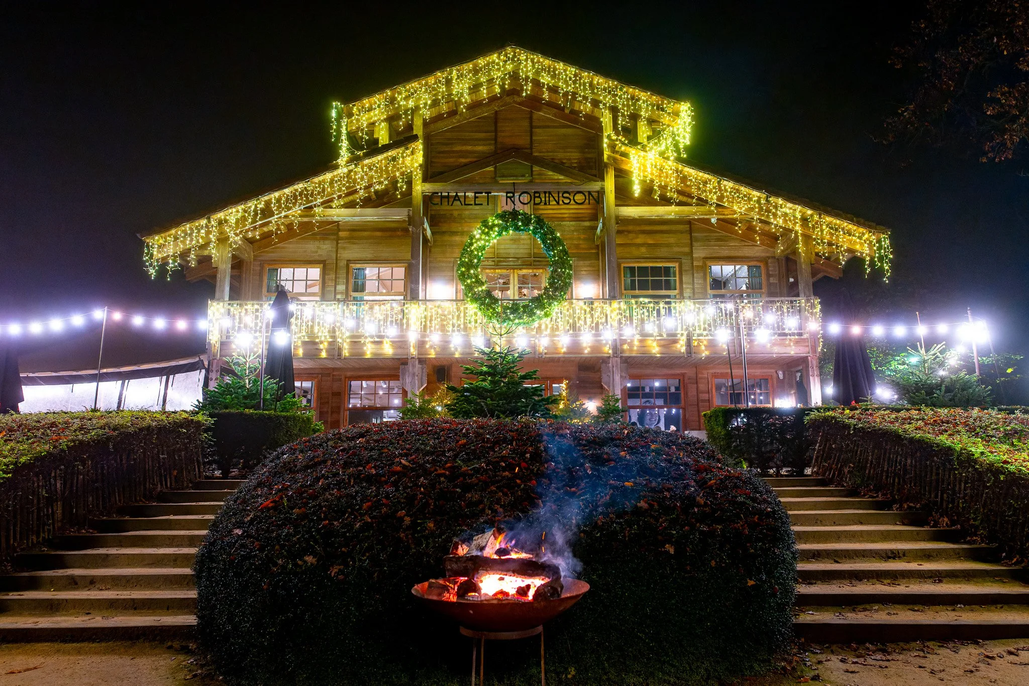 Chalet en bois décoré de lumières de Noël la nuit, avec un feu de camp au premier plan, entouré de buissons et de marches menant au bâtiment.