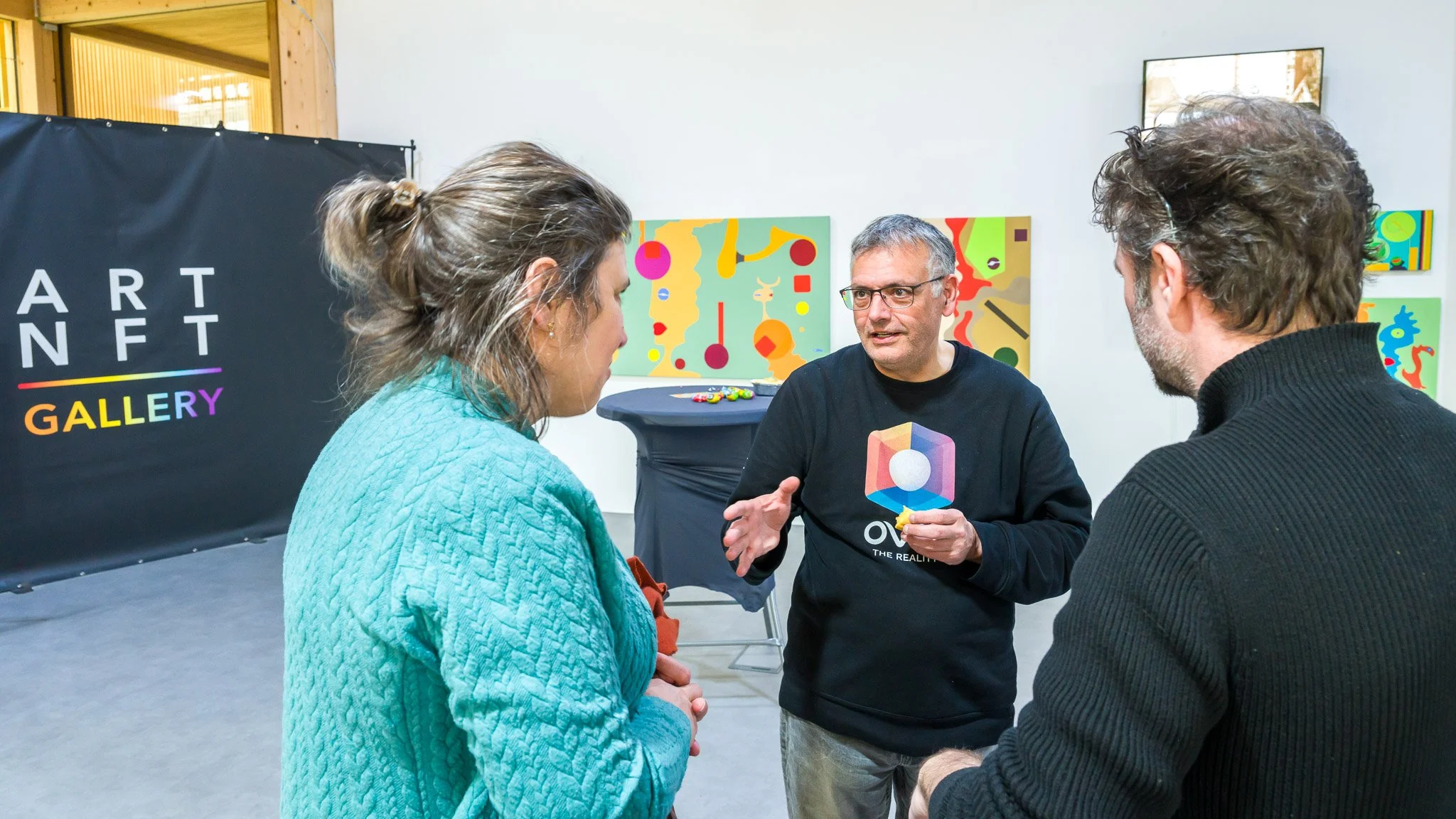 Trois personnes discutant dans une galerie d'art NFT, avec des œuvres d'art colorées en arrière-plan.