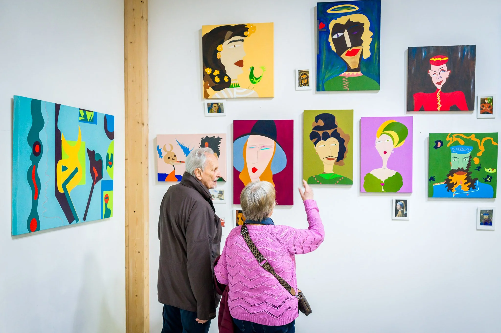 Deux personnes, un homme et une femme âgées, regardent une collection de peintures modernes colorées dans une galerie d'art. La femme pointe du doigt une œuvre.