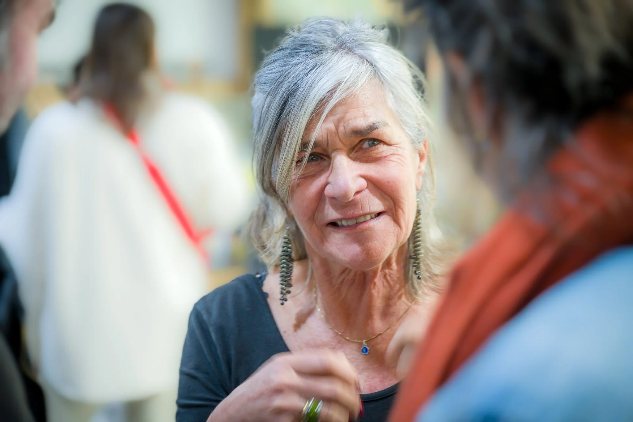 Une femme âgée souriante, portant des boucles d'oreilles pendantes et un collier, en conversation avec deux personnes floues à l'arrière-plan.