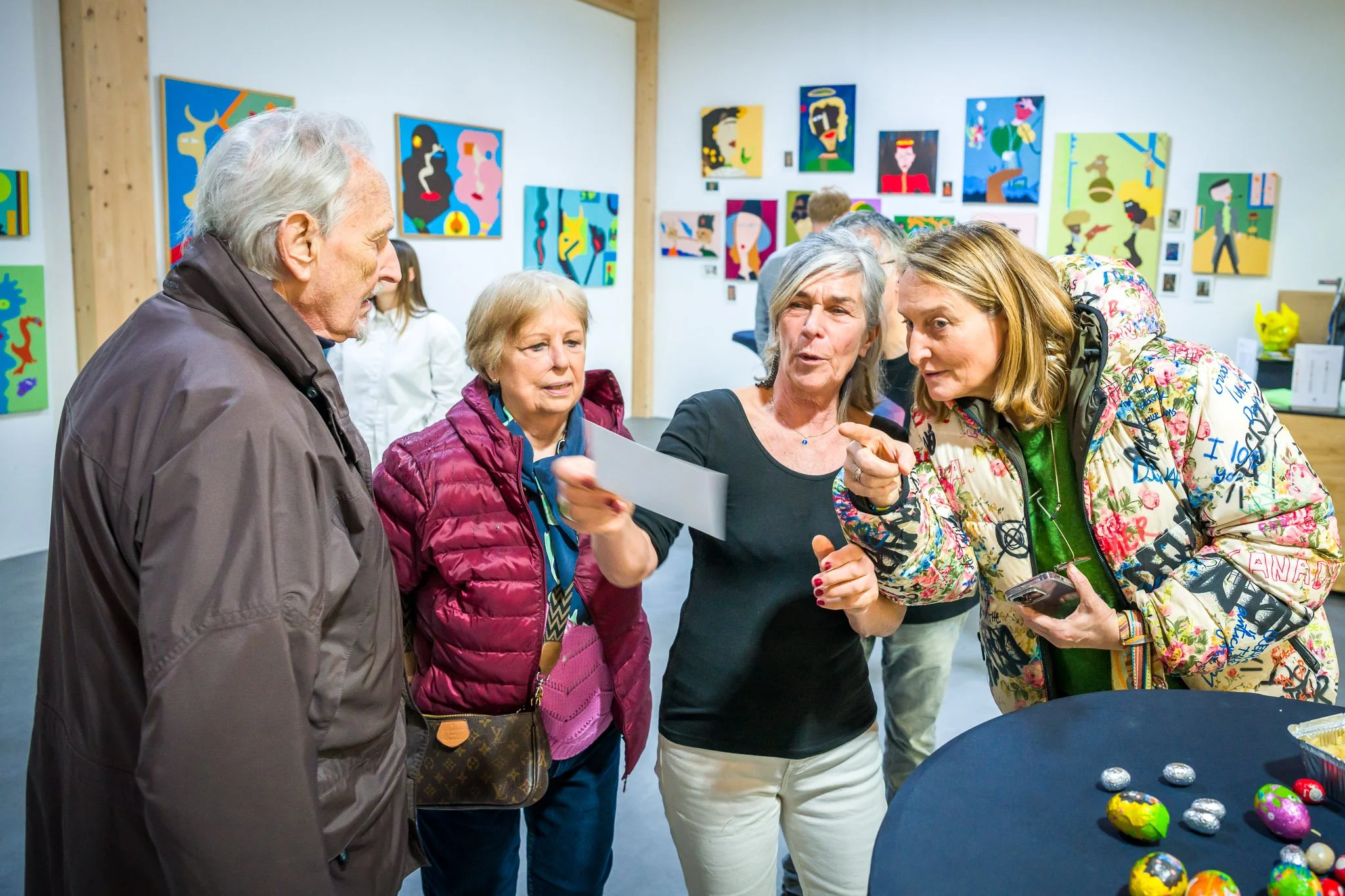 Quatre personnes discutent dans une galerie d'art, avec des peintures colorées accrochées au mur en arrière-plan.