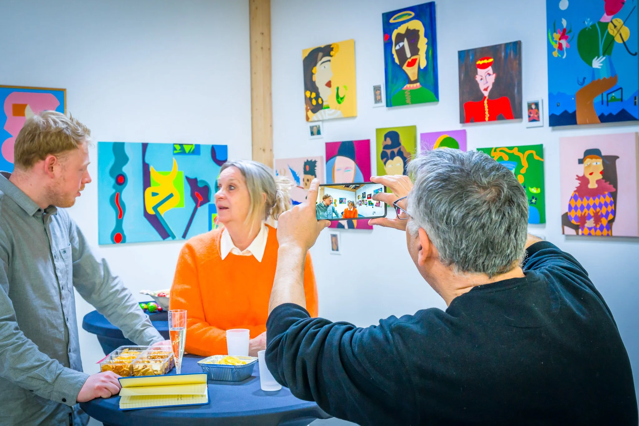 Des visiteurs échangent autour d’une table lors du vernissage de l’exposition d’Anne de Decker, avec des tableaux colorés aux murs.