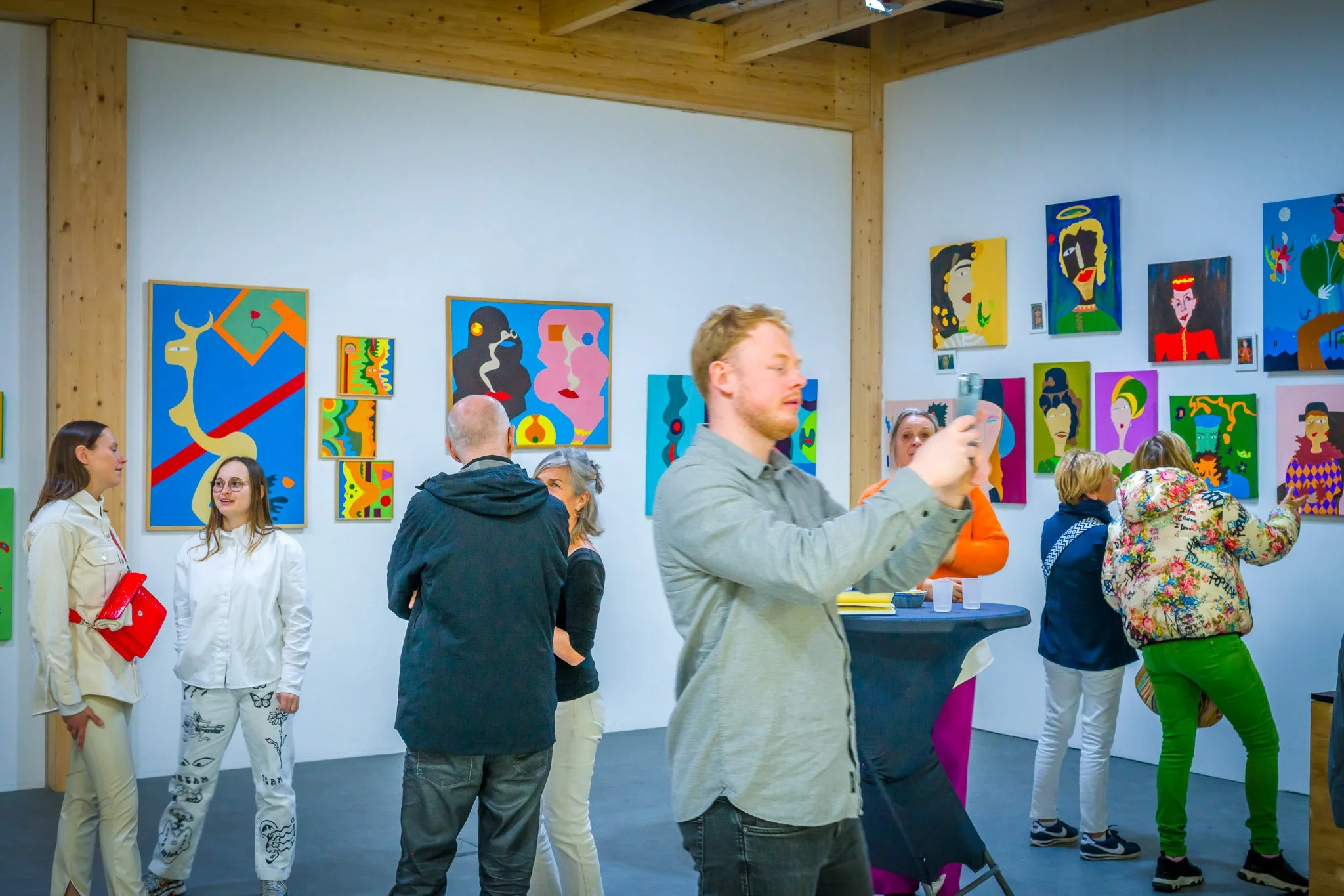 Des personnes dans une galerie d'art avec des peintures colorées sur les murs. Un homme prend une photo avec son téléphone tandis que d'autres personnes discutent ou regardent les œuvres.