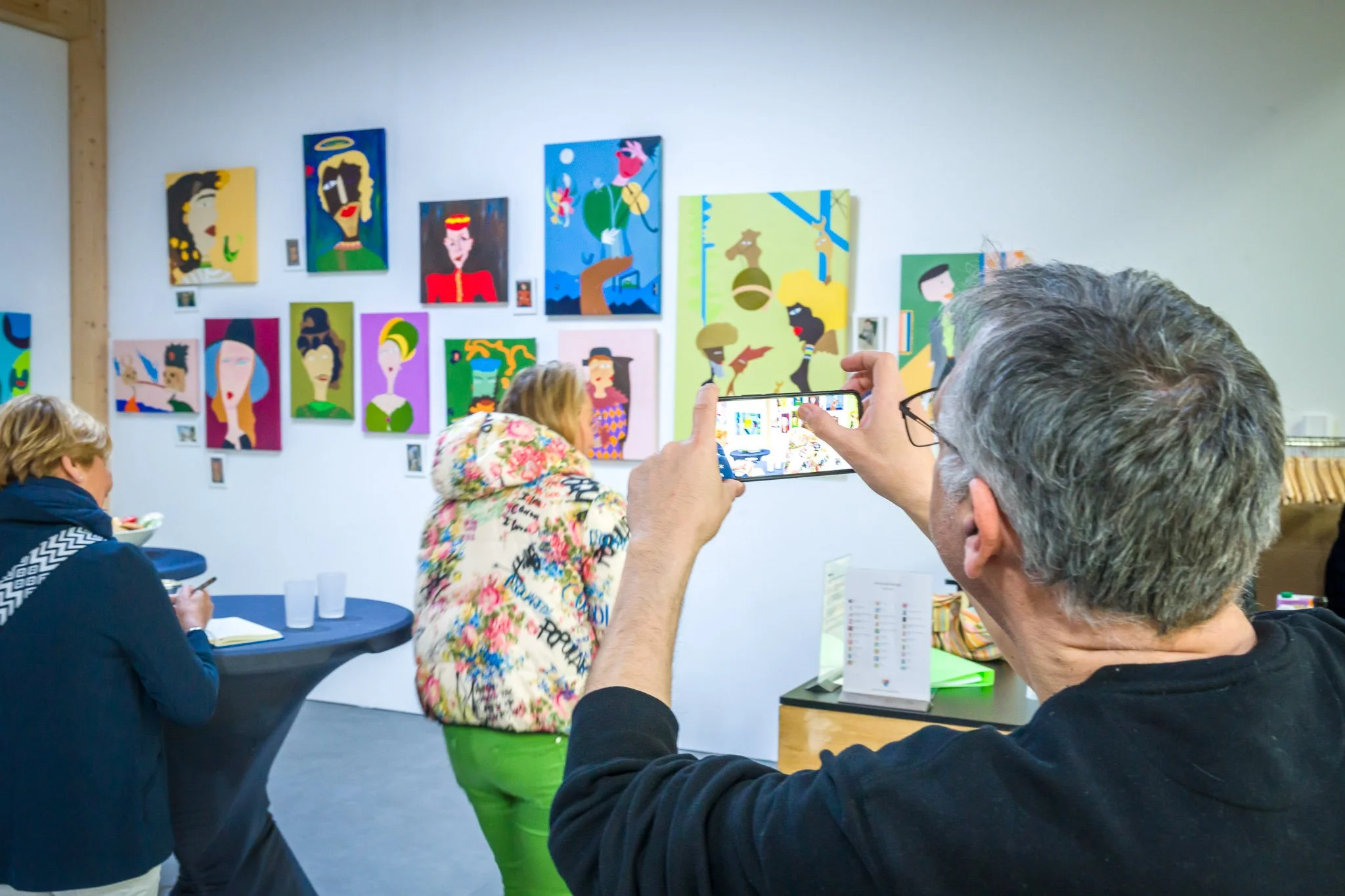Personne en train de prendre une photo d'une exposition de peintures colorées avec un téléphone, dans une galerie d'art. D'autres visiteurs sont en train de regarder les œuvres.