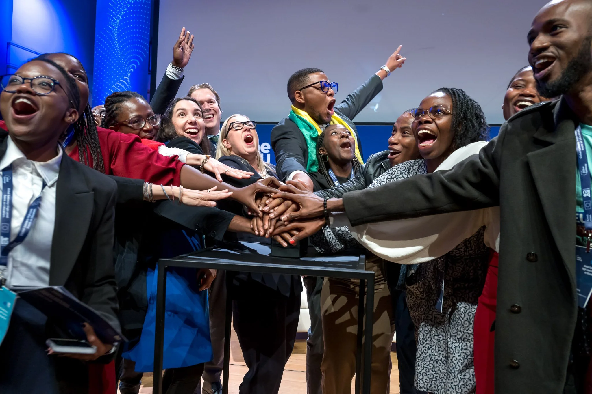 Photo professionnelle d'une conférence internationale à Bruxelles. Un groupe de personnes souriantes se tient ensemble, les mains jointes au centre, manifestant joie et enthousiasme lors d'un événement public.