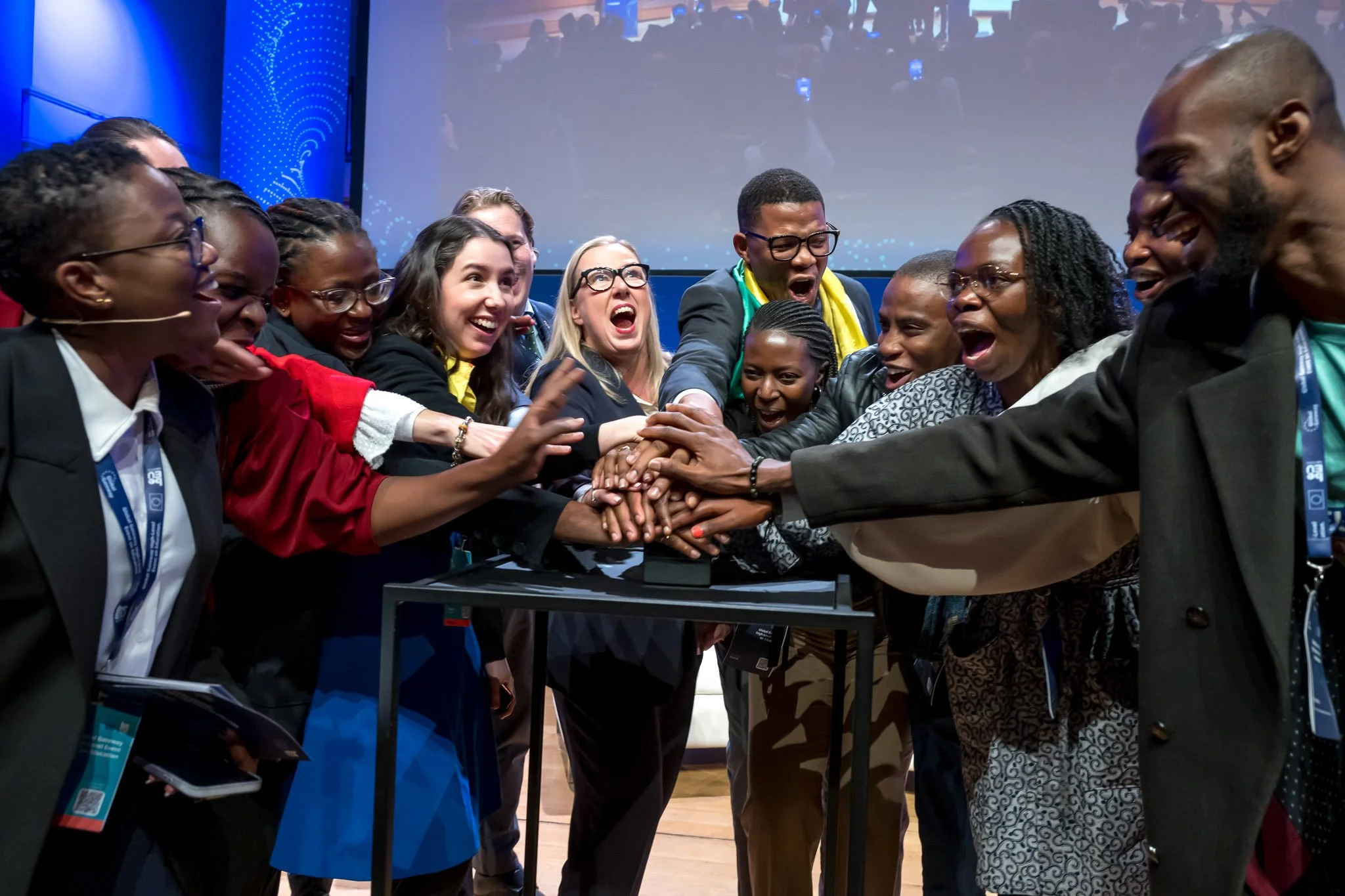 A group of smiling participants lean forward and stack their hands together at the centre of a conference stage.