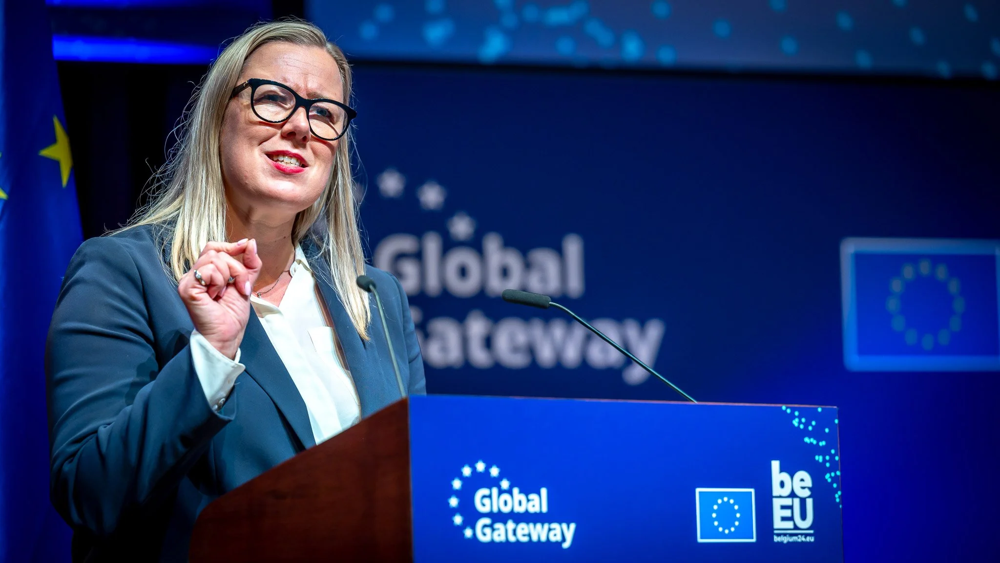 Une femme portant des lunettes, en train de parler lors d'une conférence ou d'un discours politique, avec un fond bleu portant le texte "Global Gateway" et des logos de l'Union Européenne.