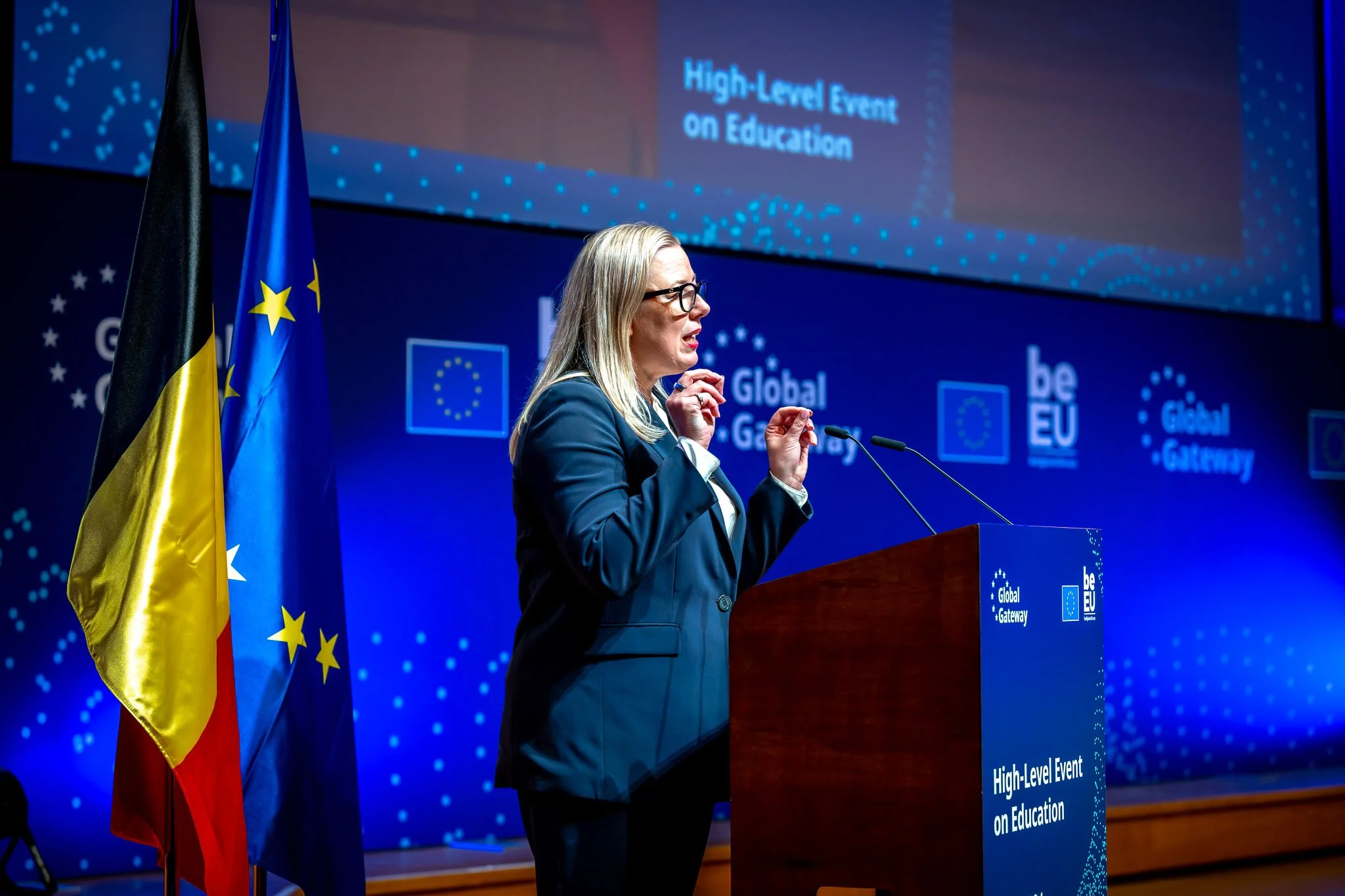 A person standing at a podium during an event on education, with European Union and Belgian flags. A blue backdrop featuring the words ‘High-Level Event on Education’ and ‘Global Gateway’.