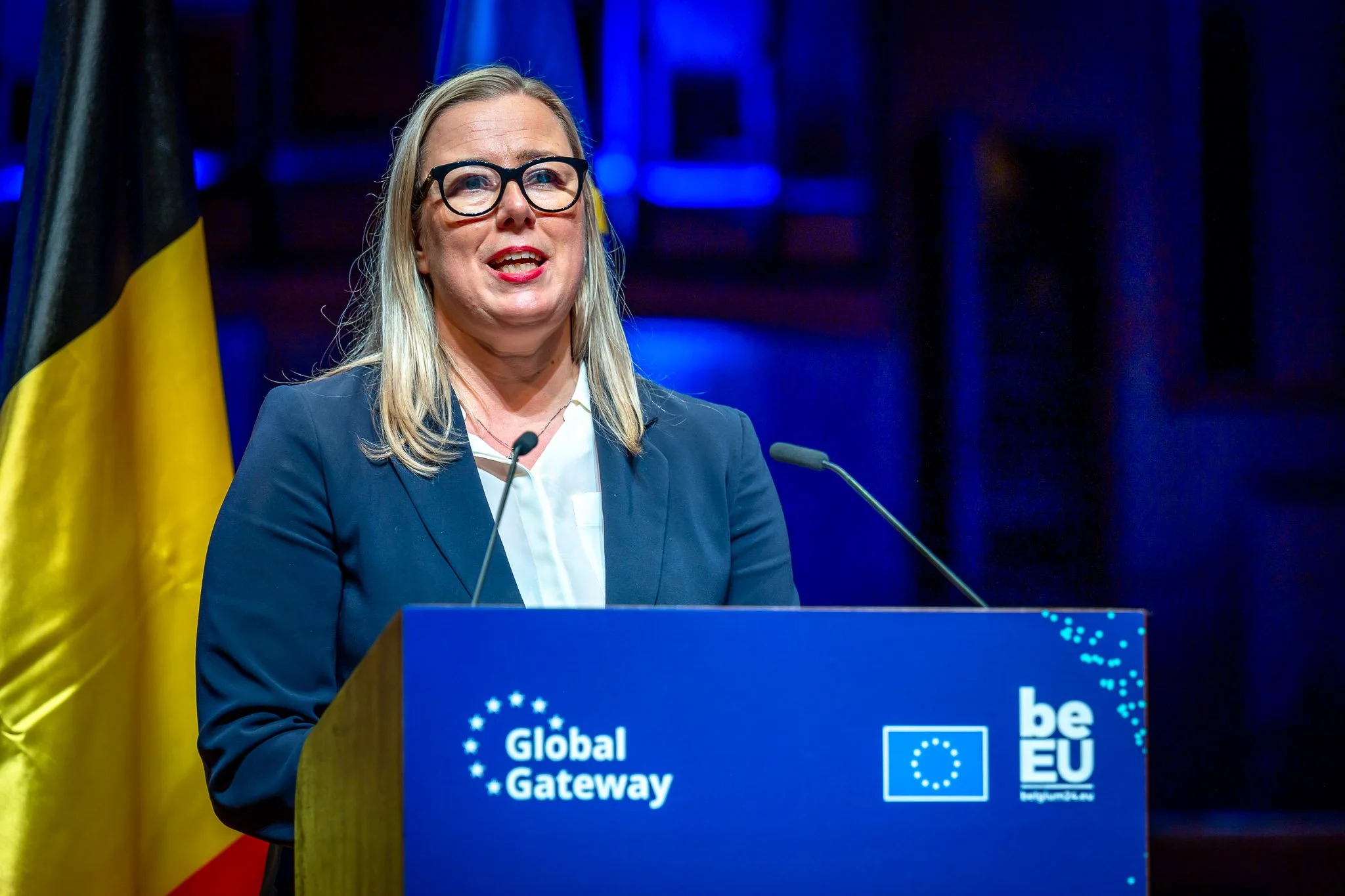 A woman speaking at a “Global Gateway” conference in front of a flag, with a lectern bearing the EU and “beEU” logos.