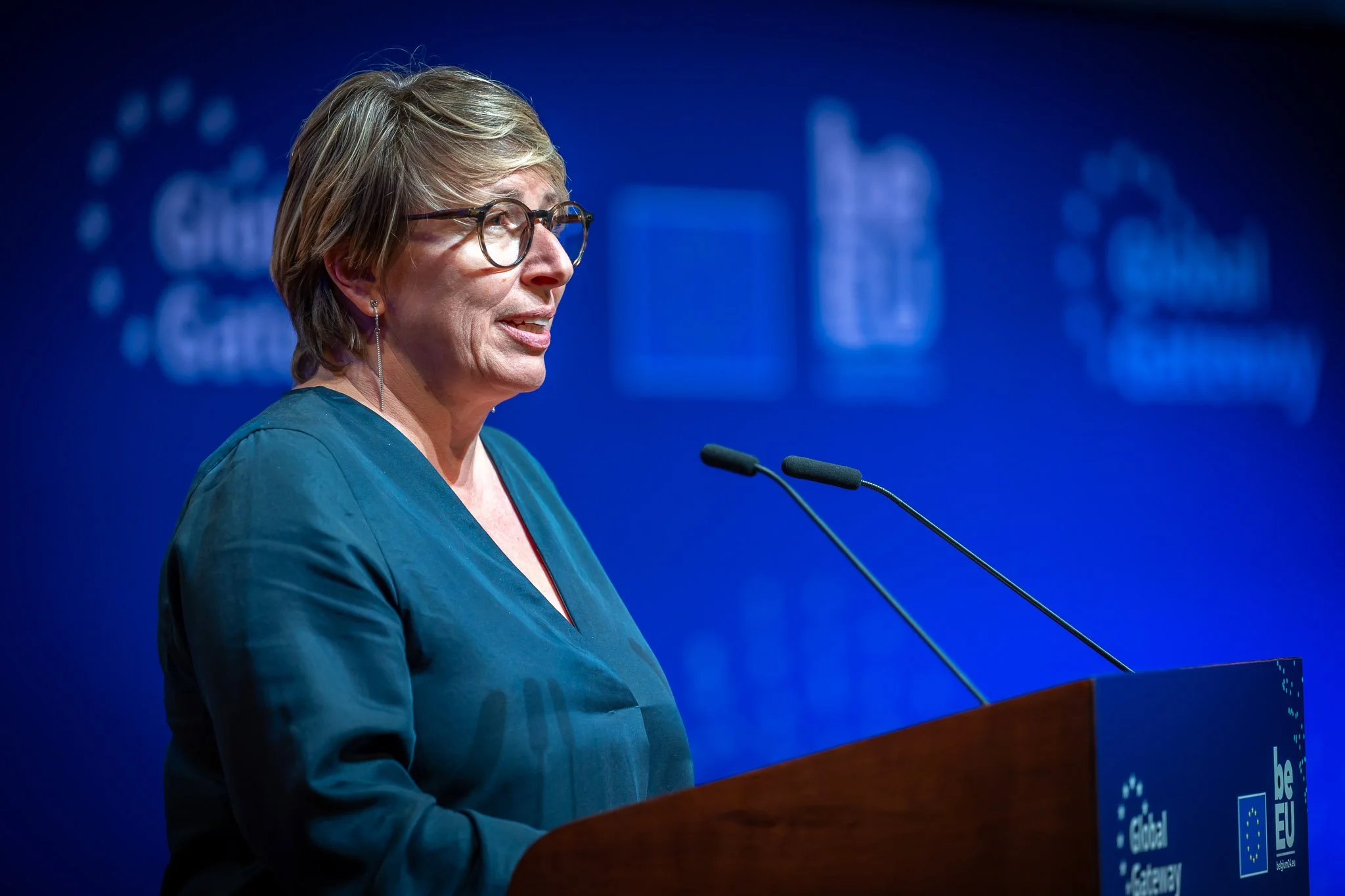 A woman giving a speech at a lectern during a ‘Global Gateway’ event. The background is blue with EU logos.