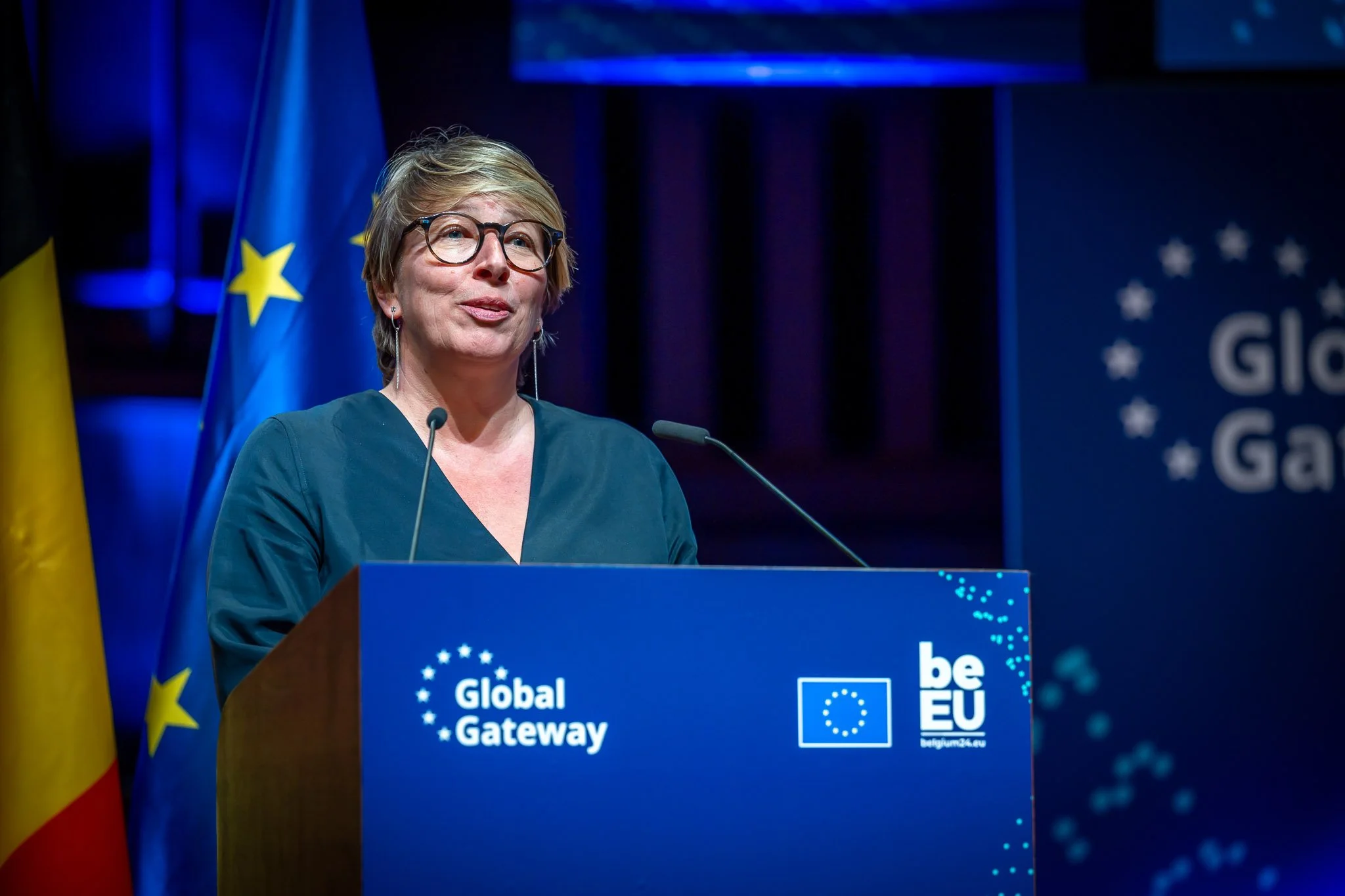 A woman is speaking at a Global Gateway event, with European Union and Belgian flags in the background.