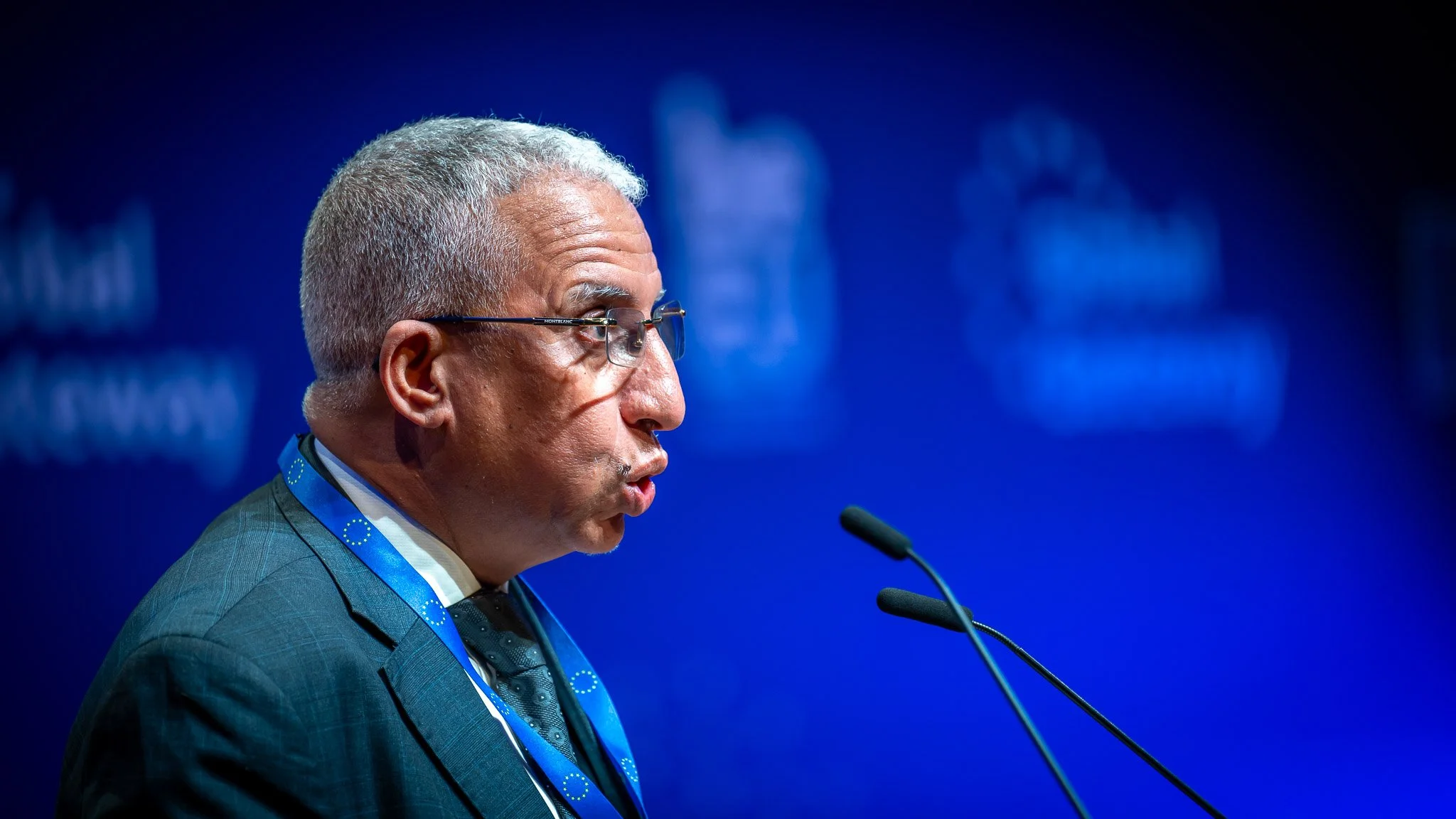 A man wearing glasses is speaking into a microphone at an event, against a blue background with a blurred logo.