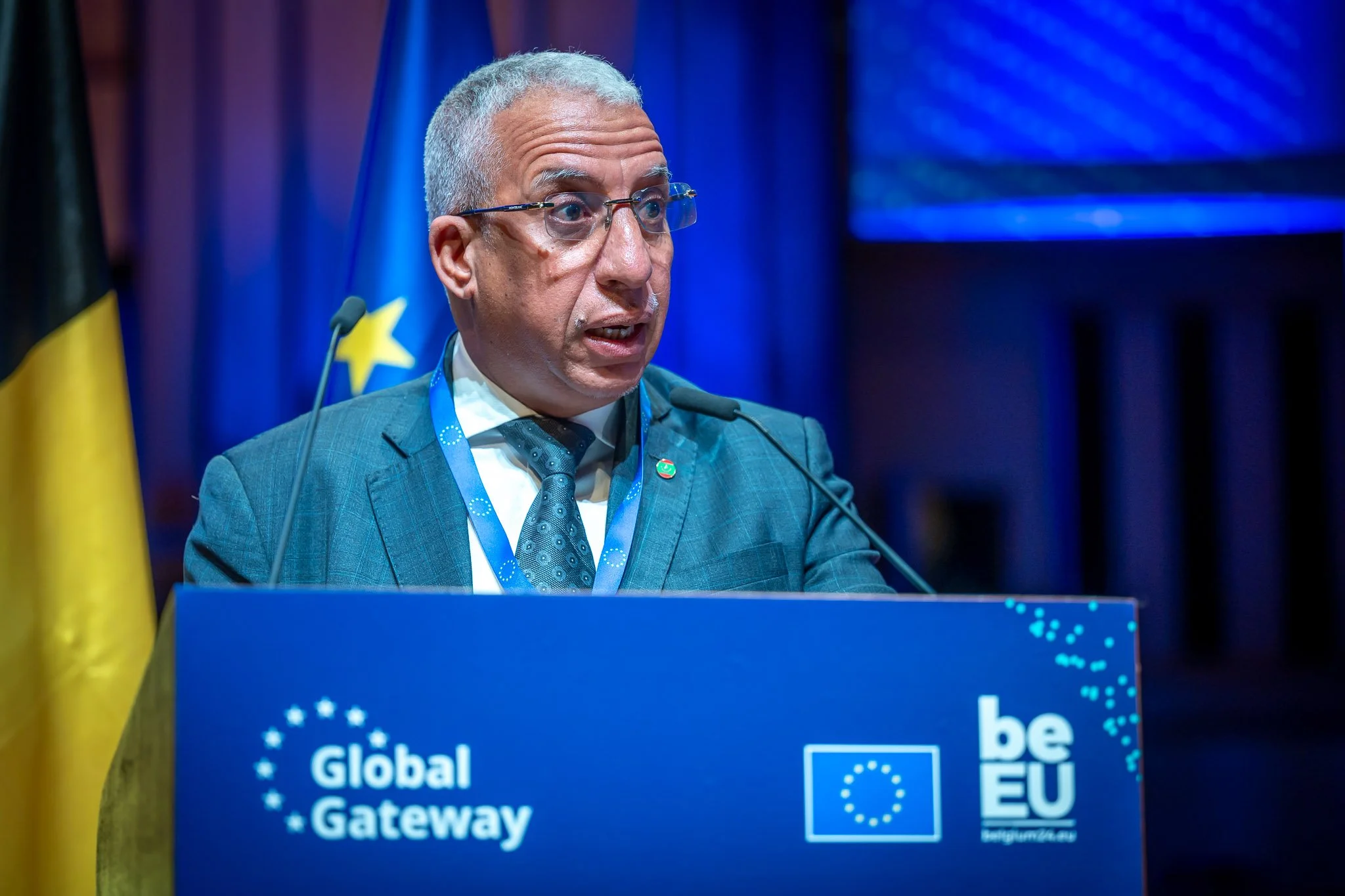 A man in a suit speaking at a ‘Global Gateway’ lectern, with flags in the background.