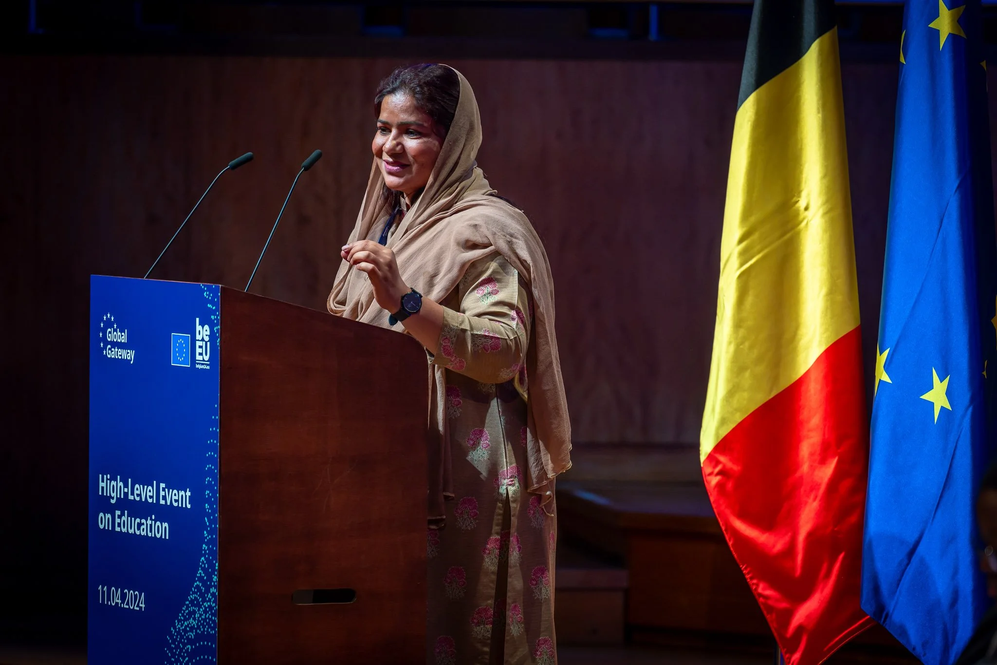 A woman speaking at an event on education, with the flags of Belgium and the European Union beside her.