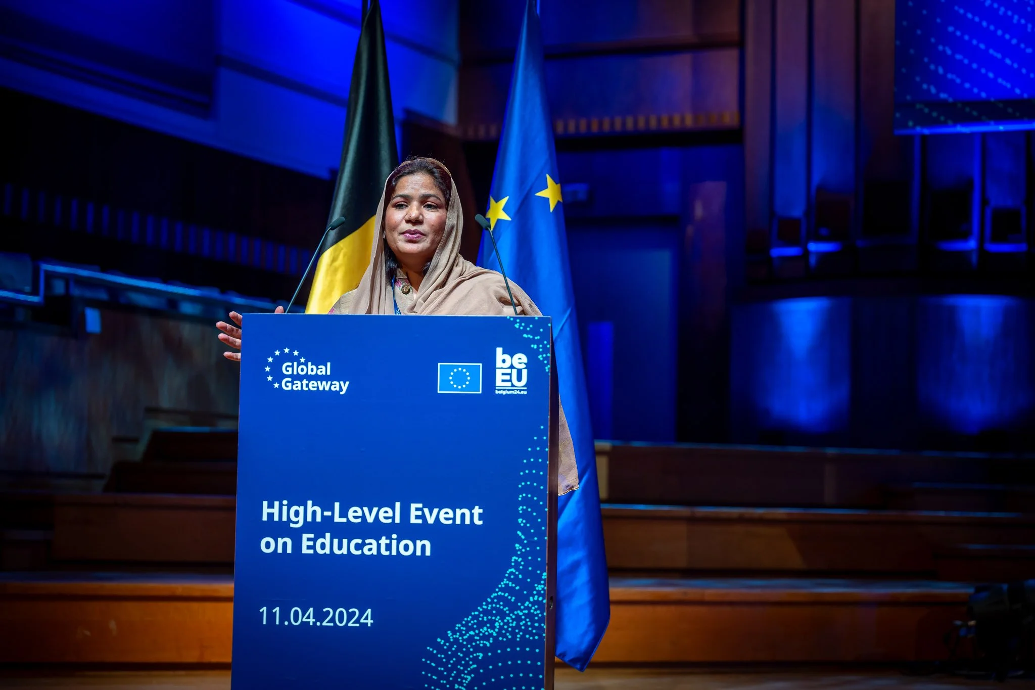 A woman is speaking at a ‘High-Level Event on Education’, with flags in the background. She is standing in front of a blue lectern bearing the Global Gateway and beEU logos, dated 11 April 2024. The atmosphere is bright, with a blue light