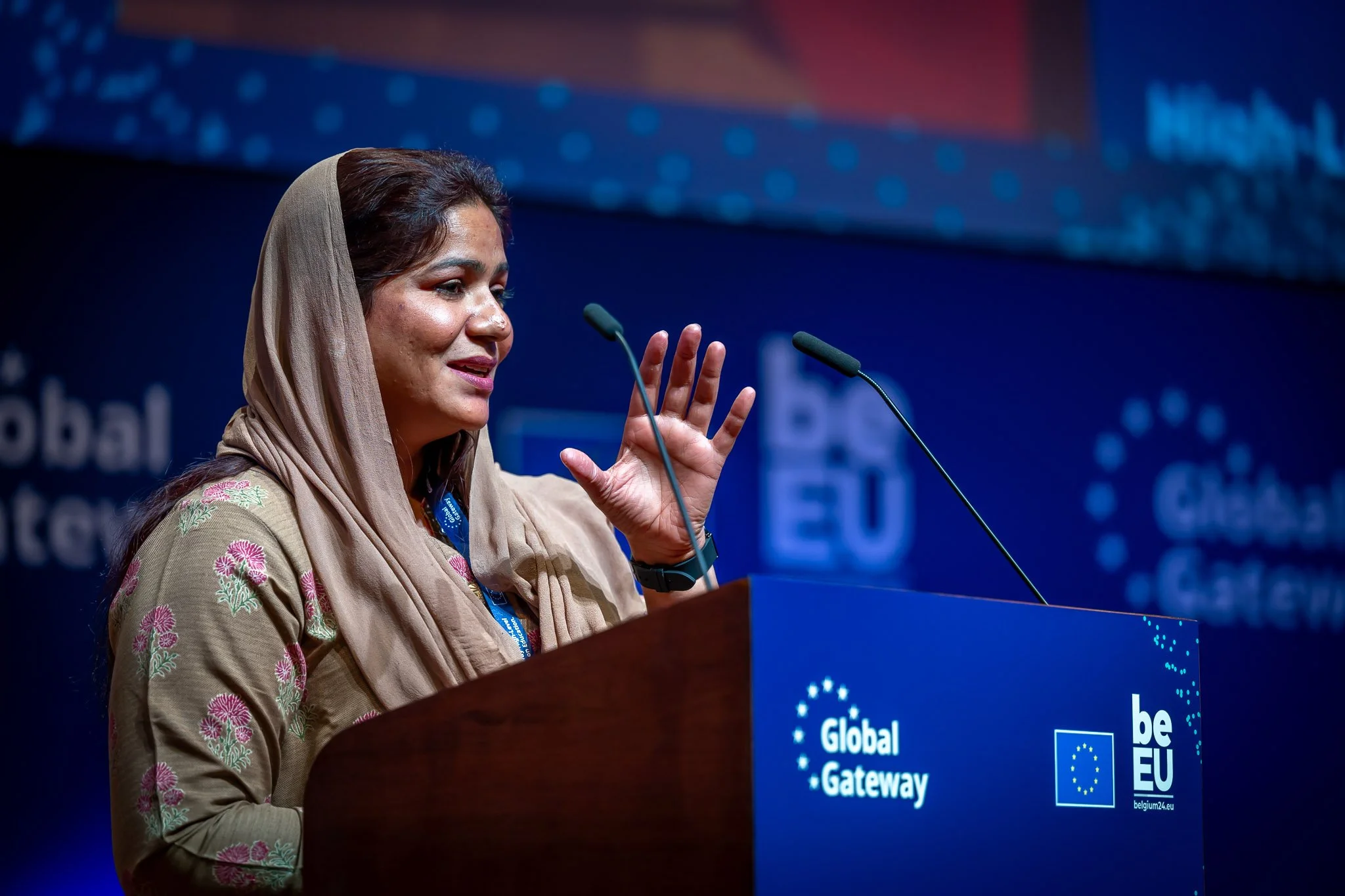 A woman in traditional dress is speaking on stage at a lectern marked ‘Global Gateway’. The background is blue with the event’s logos. She is gesturing with her hand as she speaks.