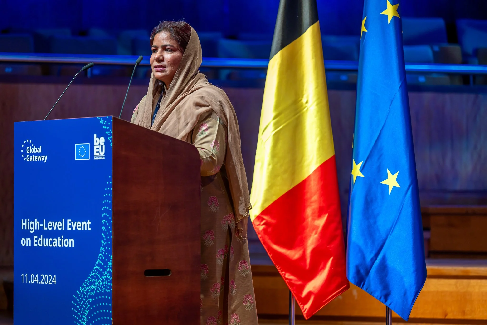 A woman is speaking at an international educational event, standing behind a lectern with the Belgian and European flags.