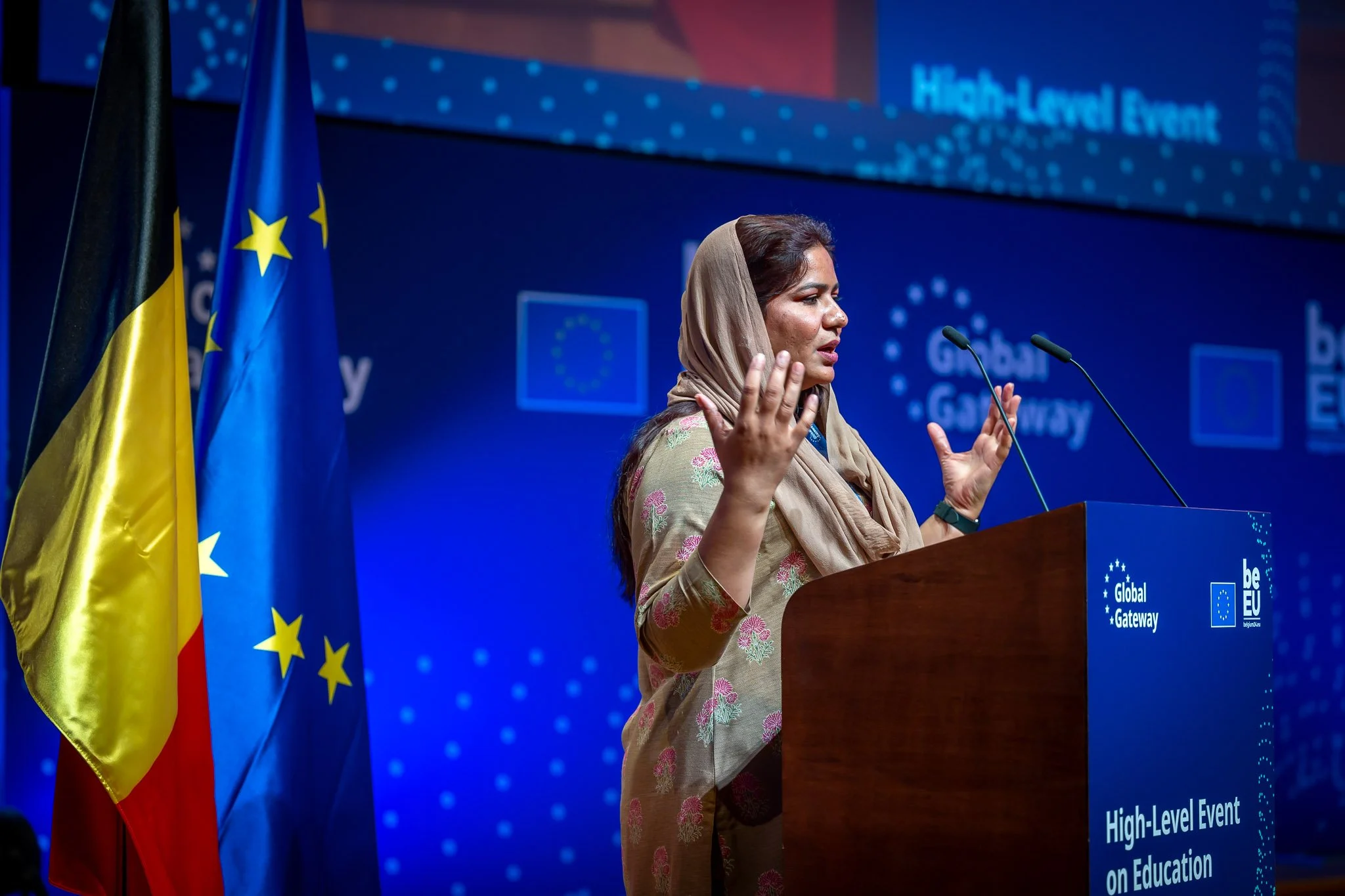 A person speaking at an event on education, in front of European Union and Belgian flags, with the ‘Global Gateway’ logo in the background.