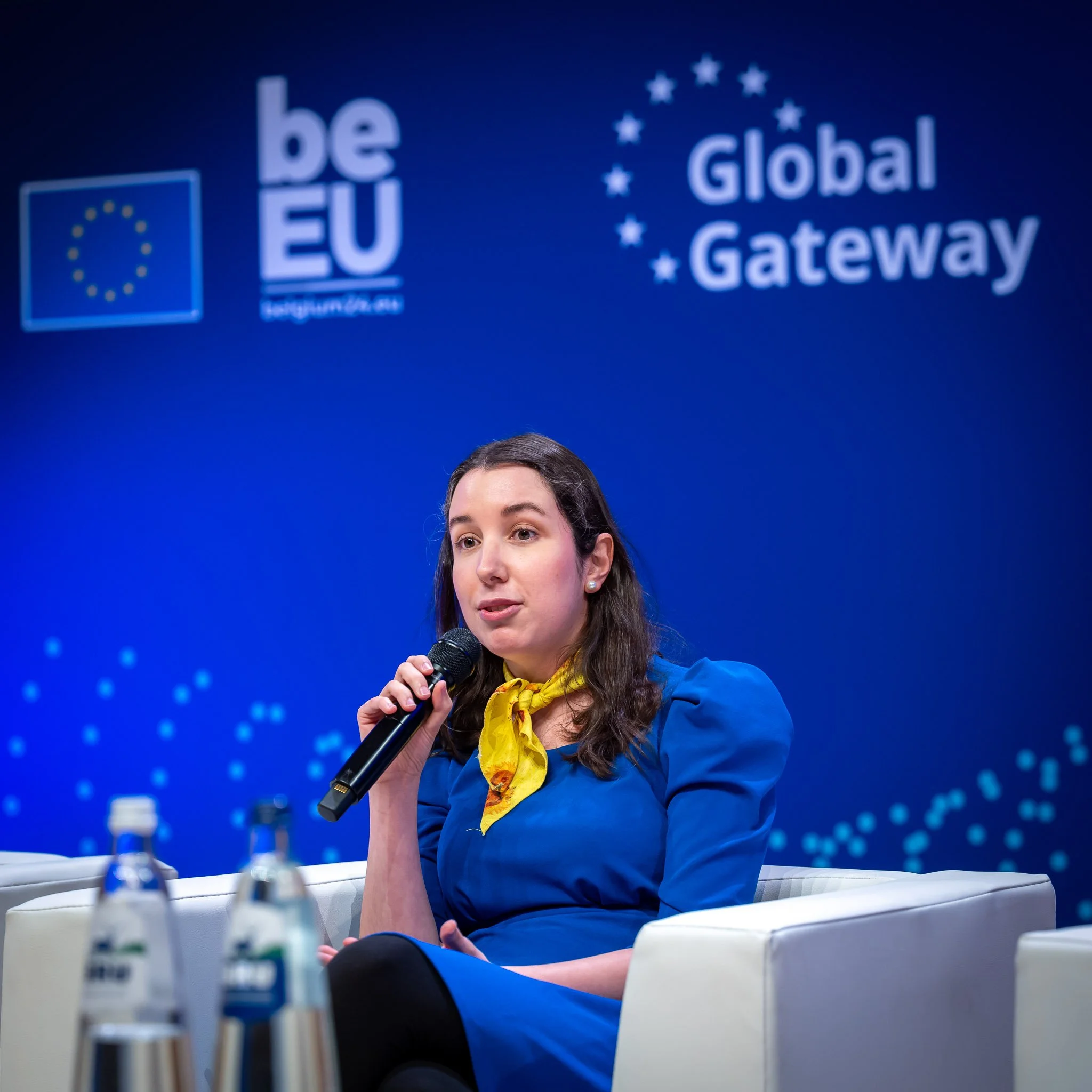 A person speaking into a microphone on a stage with the ‘Global Gateway’ and ‘beEU’ logos in the background. They are sitting in a white armchair and wearing a blue dress with a yellow scarf. Two bottles of water are visible in the foreground.