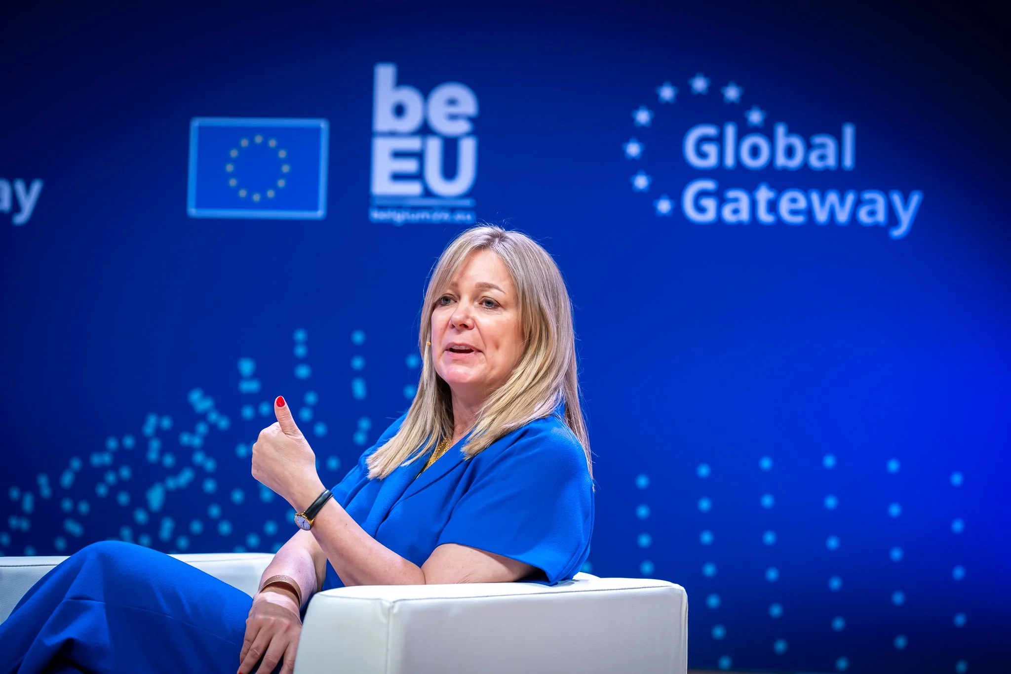 A woman in a blue dress is speaking at an event called “Global Gateway”, with European logos in the background.