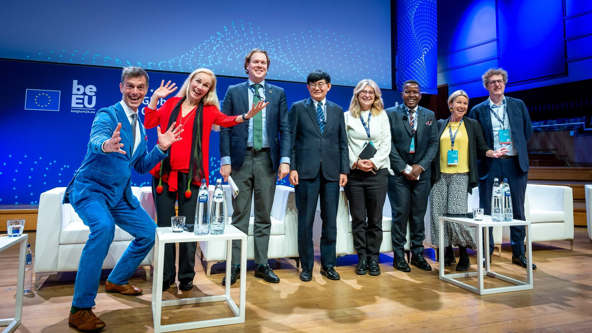 A group of participants pose together on a blue conference stage behind small tables and water bottles.