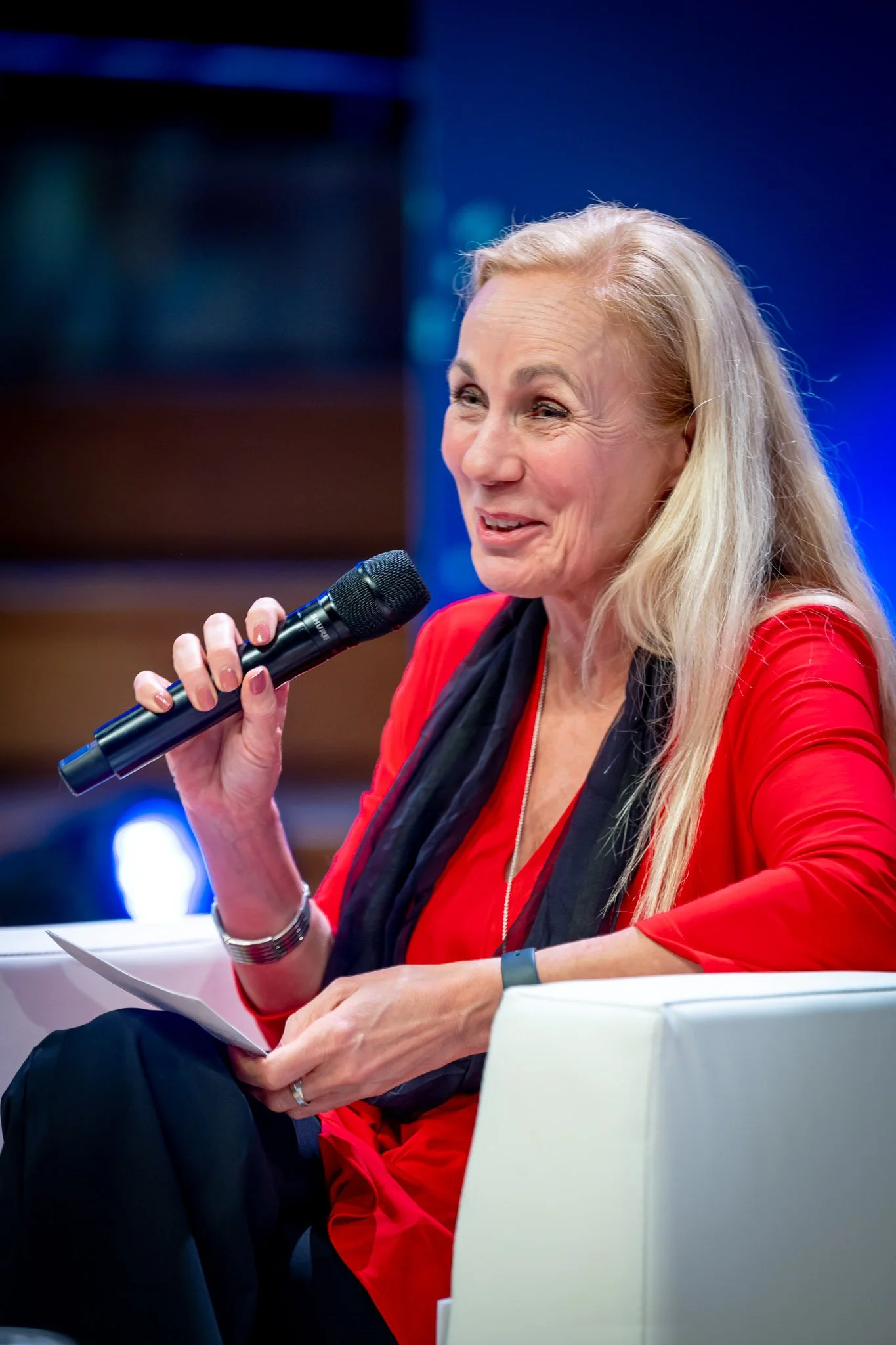 A smiling woman in a red dress holding a microphone at an event, sitting on a white sofa.