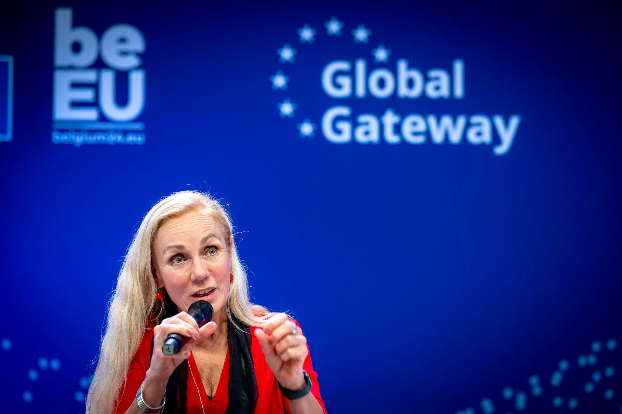 A woman holding a microphone, against a blue background with the words "Global Gateway" and "be EU" displayed.