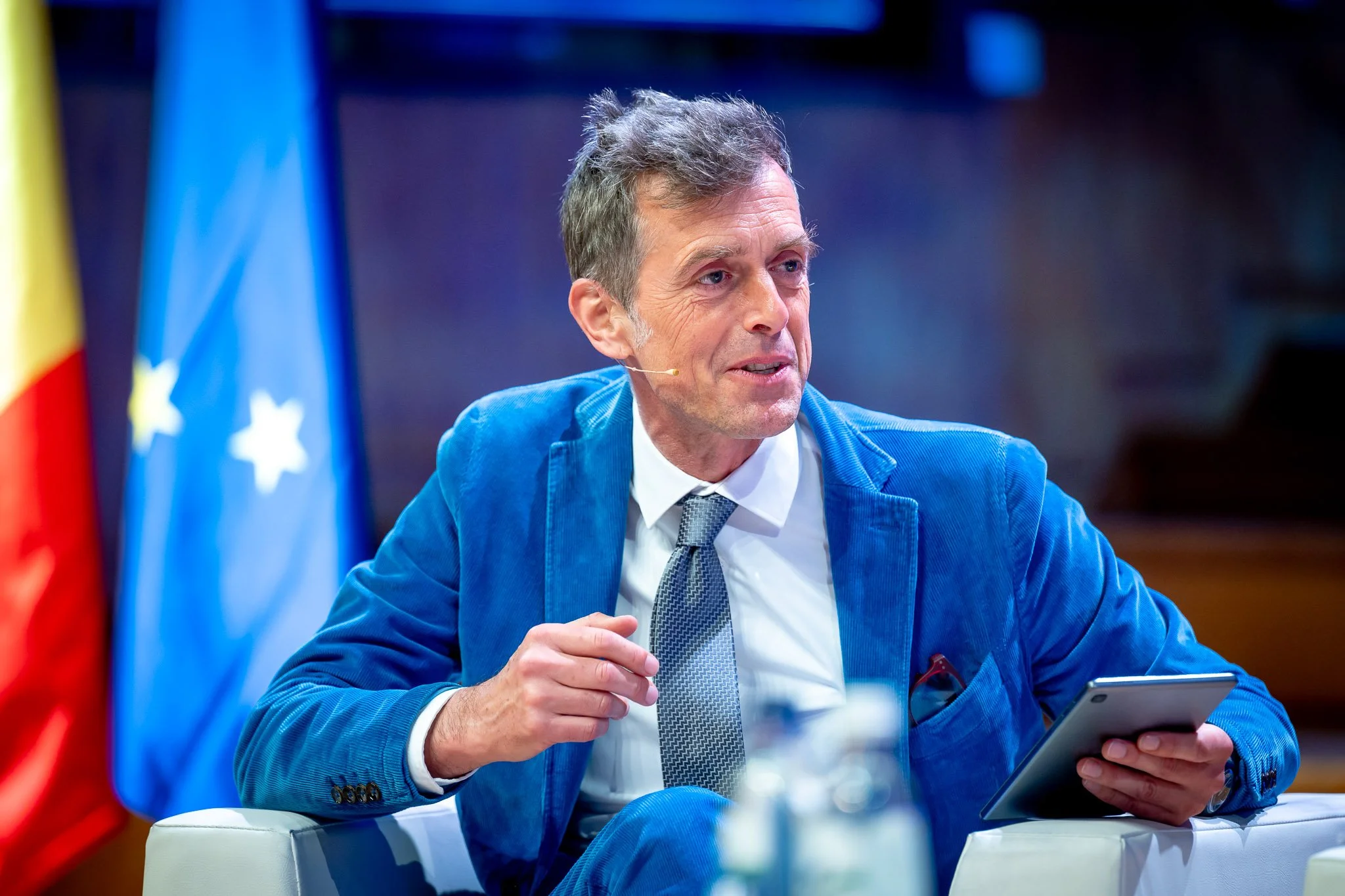 A man in a blue suit holding a tablet begins a lecture, with the European flag in the background.