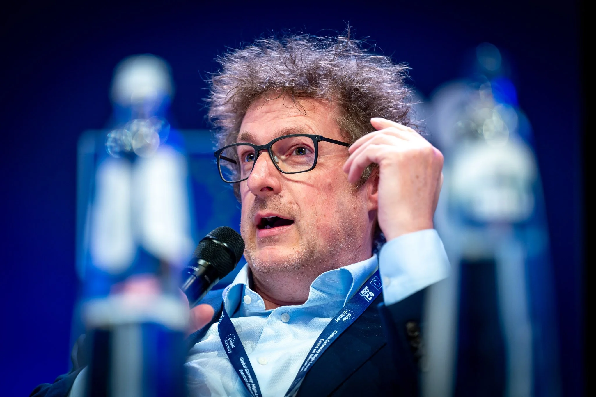 Homme avec des lunettes, parlant dans un microphone lors d'une conférence, entouré de bouteilles d'eau.