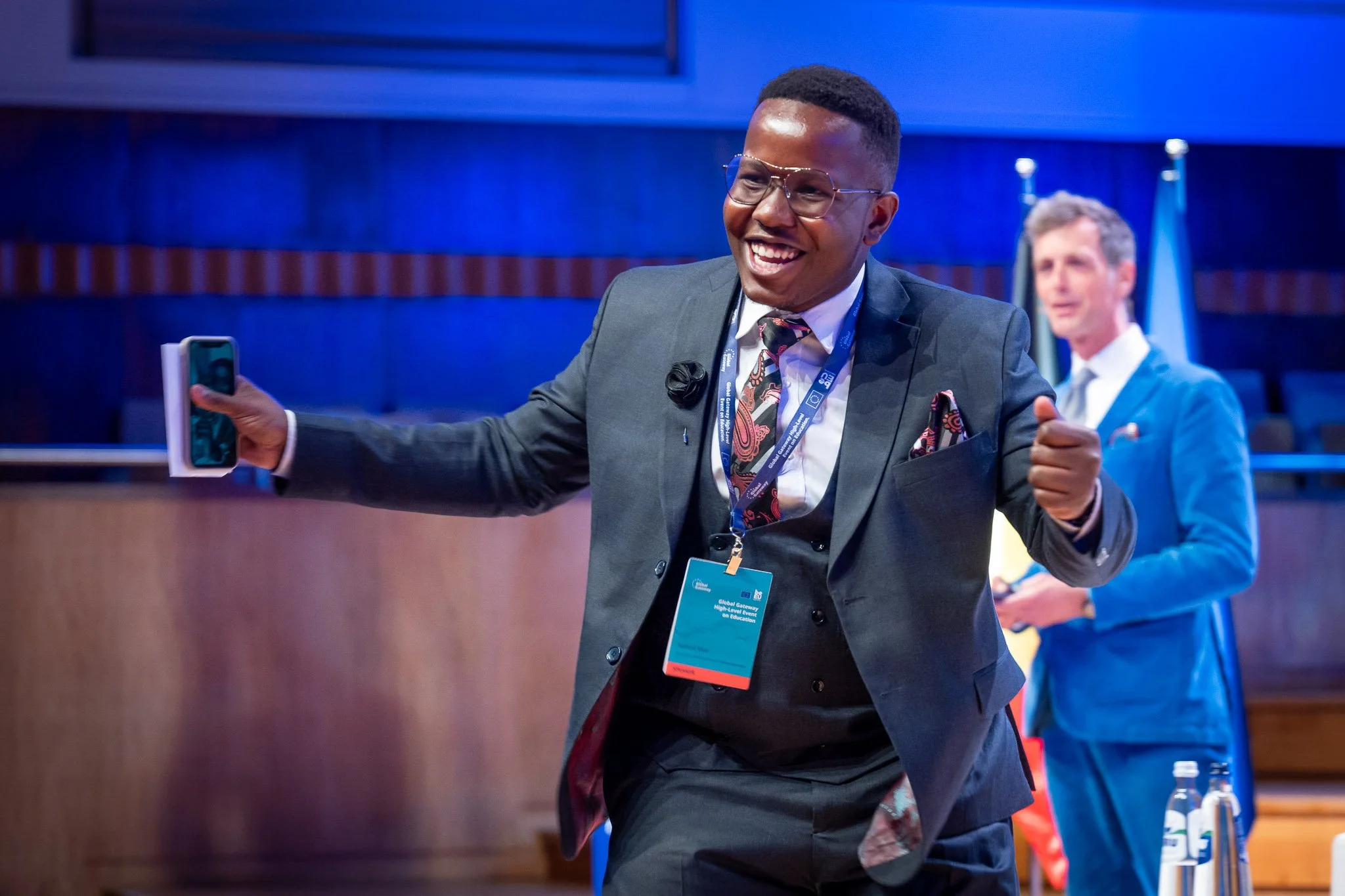 A smiling man in a suit, wearing a badge and holding a mobile phone, on a stage; another man, blurred, in the background.