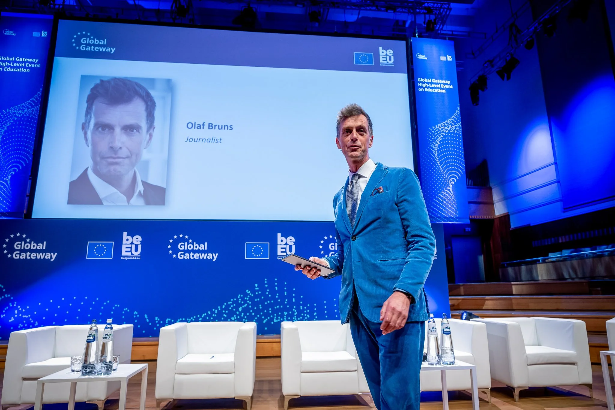 A man in a blue suit is standing on stage at a Global Gateway event. In the background, there is a screen displaying the name ‘Olaf Bruns’ and the word ‘Journalist’. The stage is set up with white chairs and bottles of water on a table