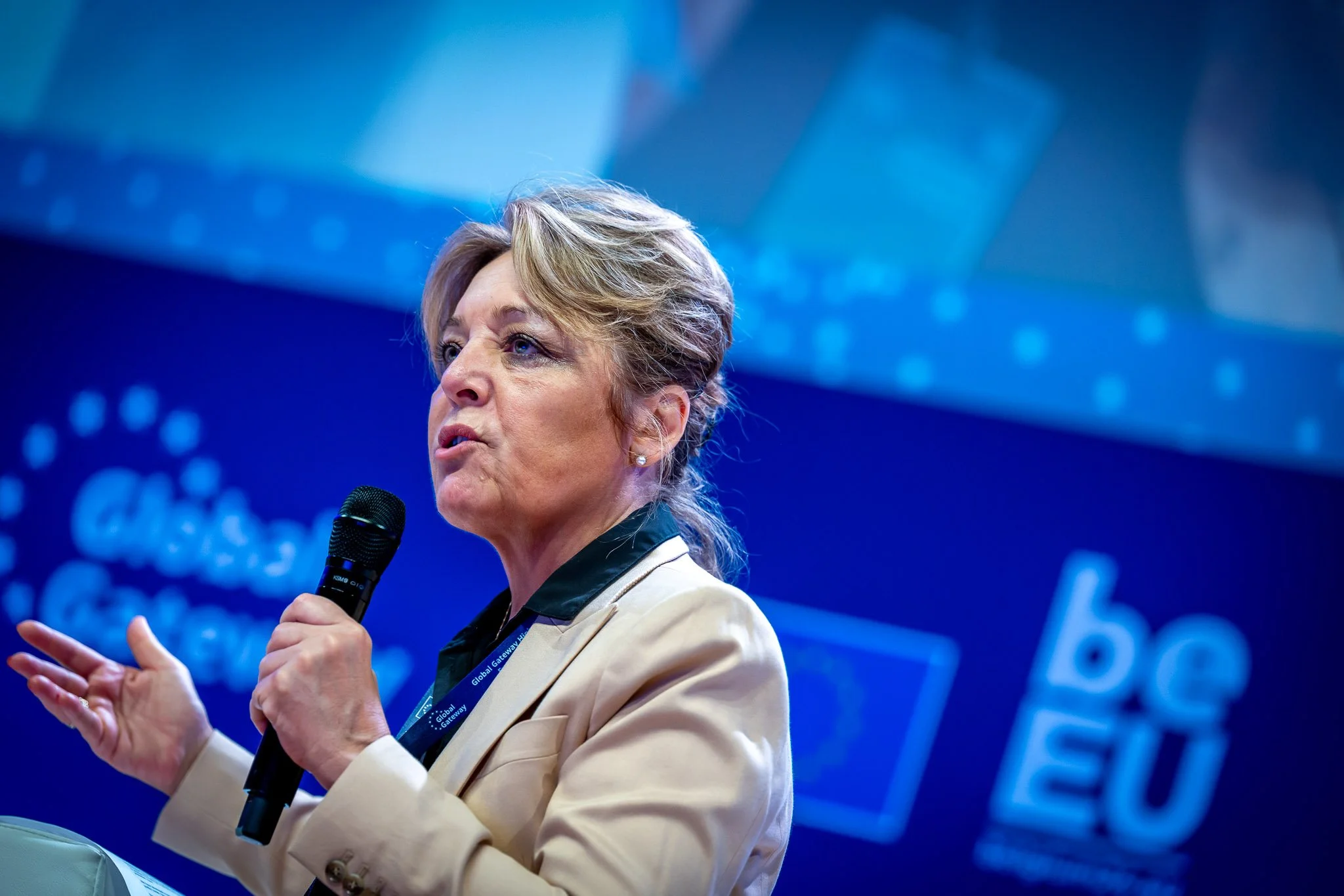 A woman holding a microphone at a conference, with a blue background and the words ‘Global Gateway’ and ‘be EU’ visible.