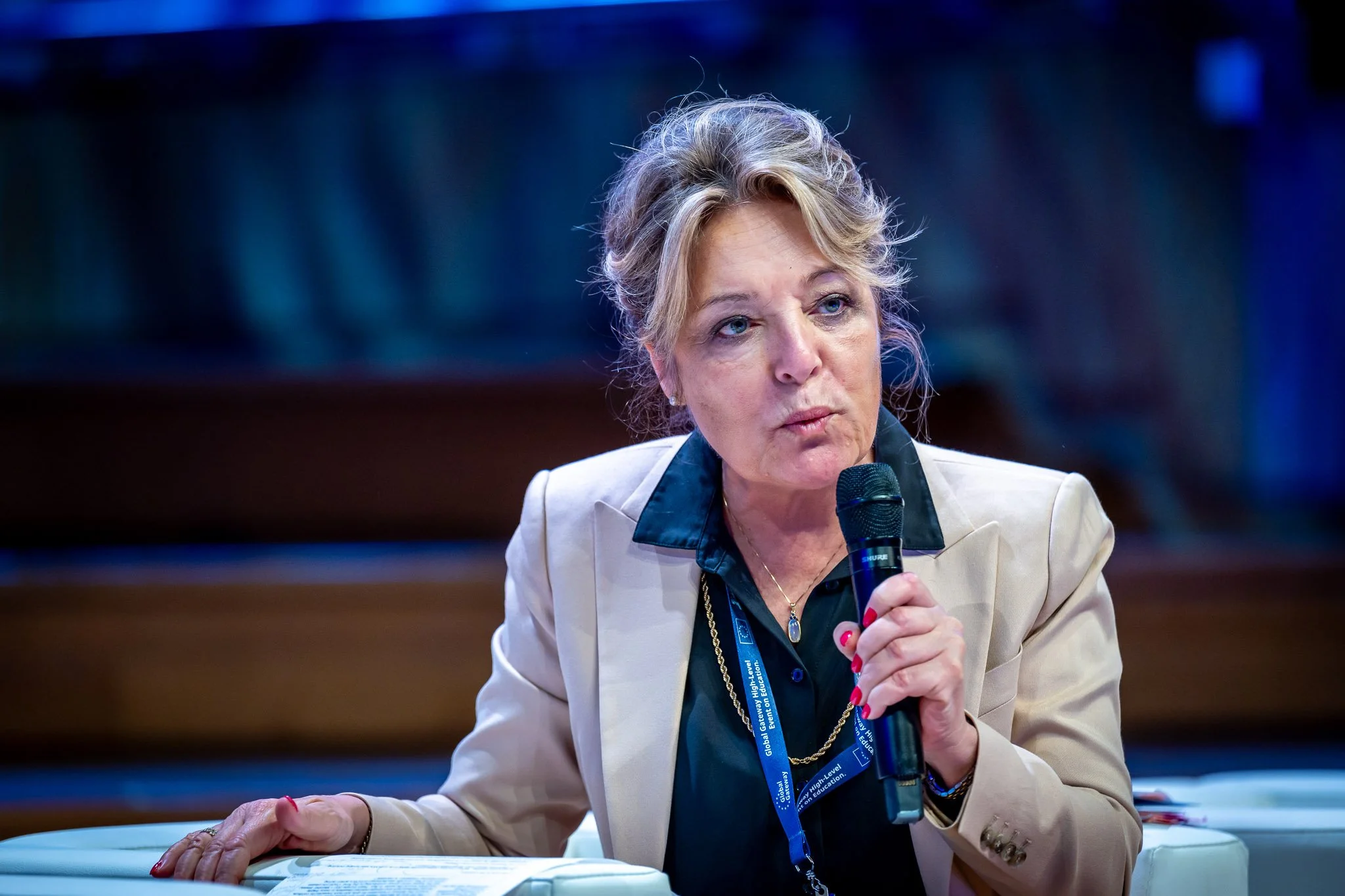 A woman in a beige suit speaking into a microphone at an event.