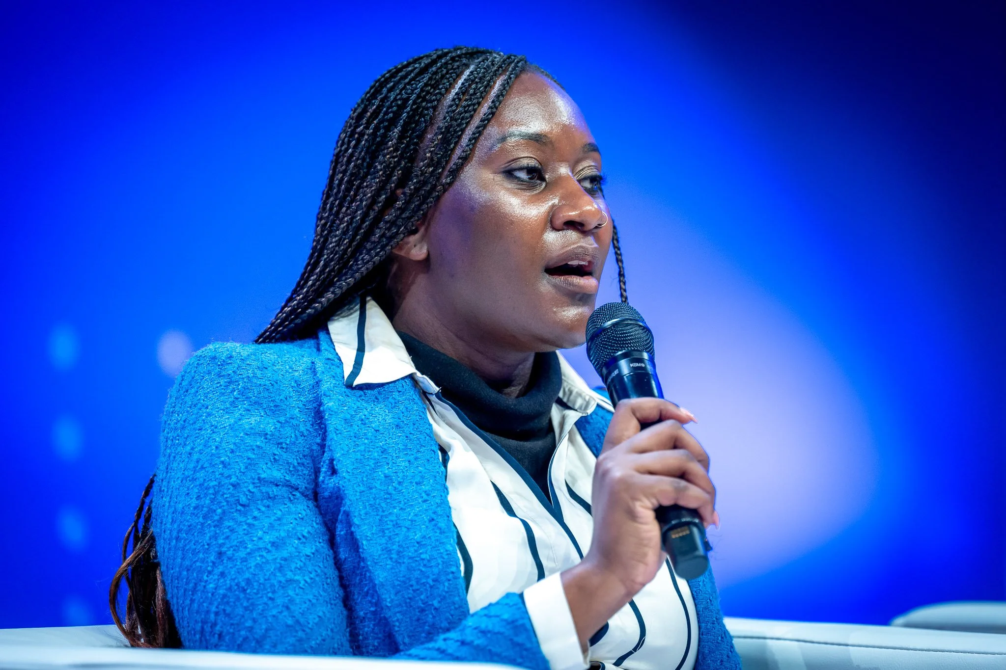 A woman speaking into a microphone at a conference, wearing a blue jacket against a blue background.