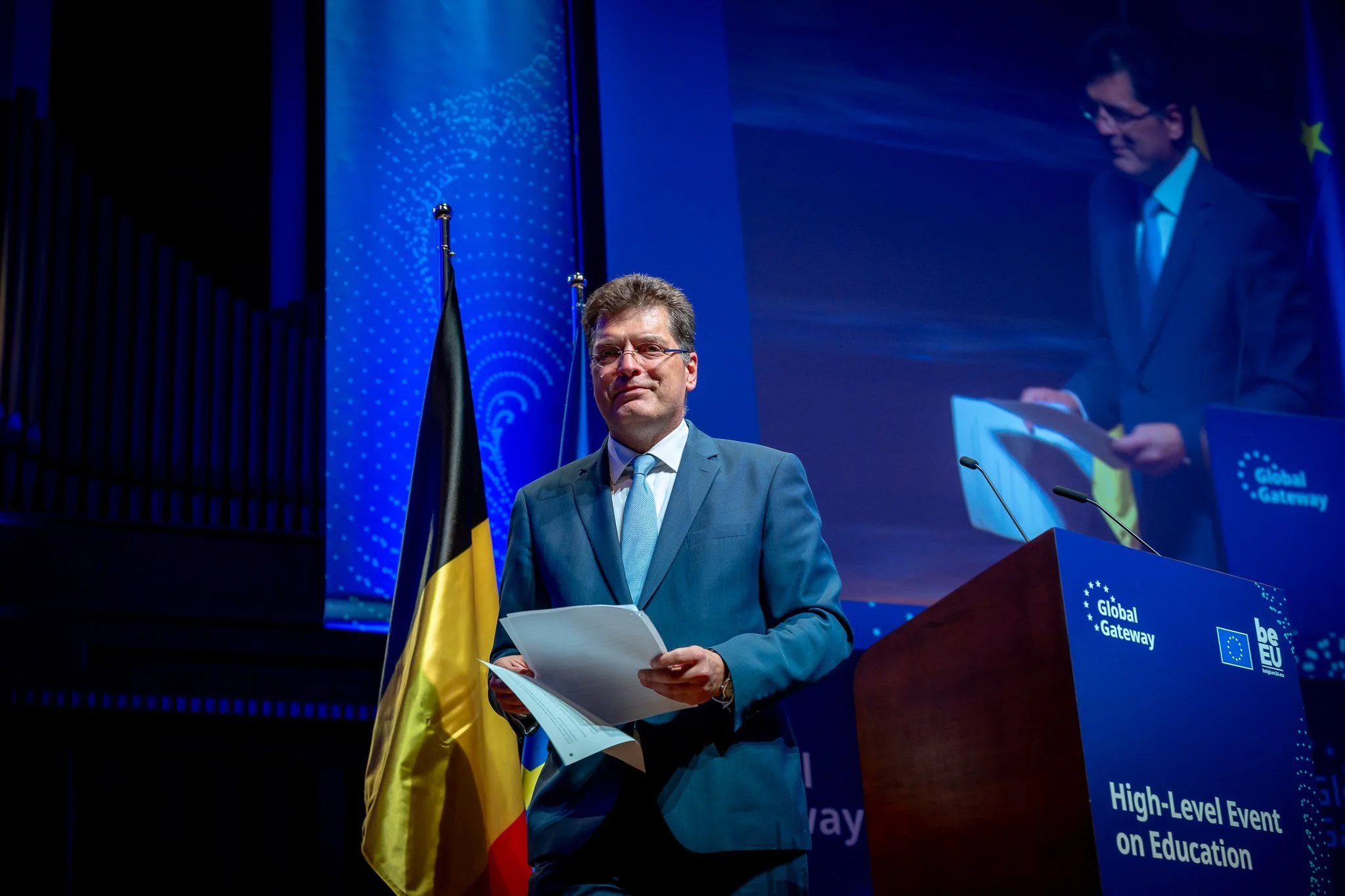 A man in a suit is holding documents on stage in front of a lectern marked ‘Global Gateway’ and ‘High-Level Event on Education’, with flags and a projection screen in the background.