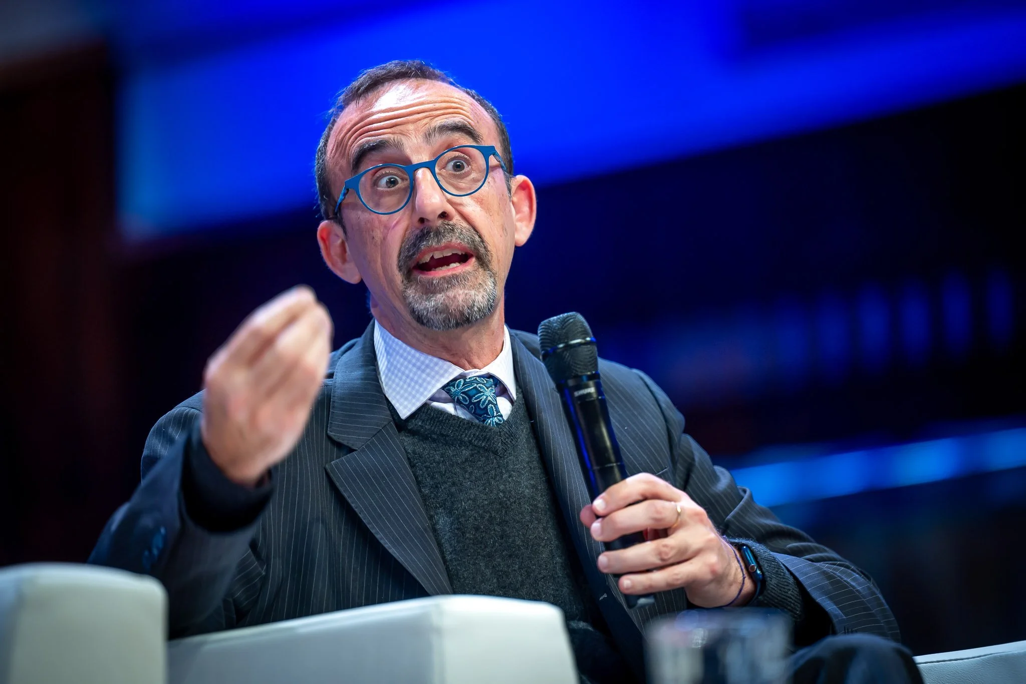 Un homme avec des lunettes bleues, tenant un micro, parle lors d'une conférence ou d'un débat.