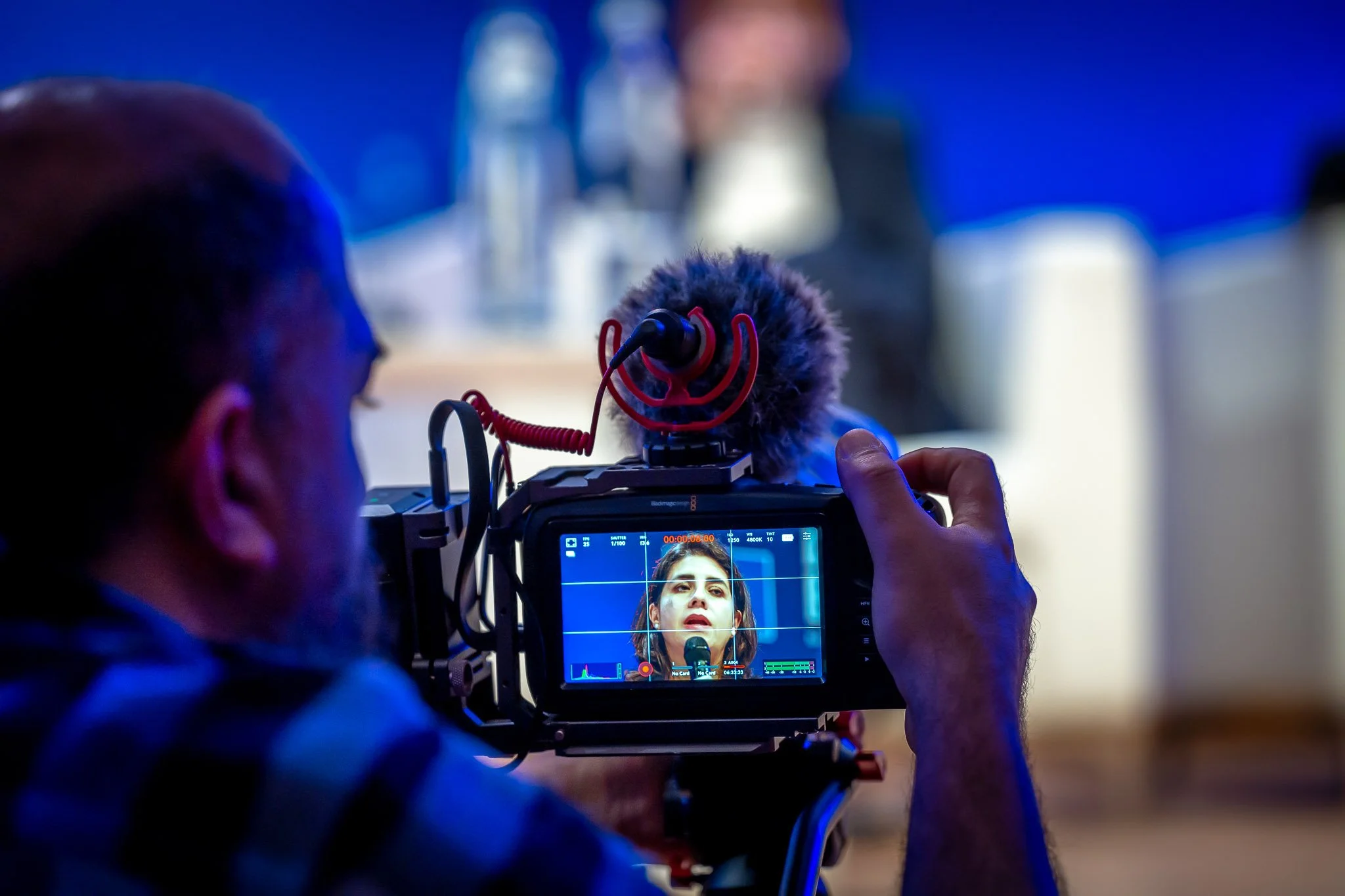 A cameraman is filming a woman speaking into a microphone, with a screen showing her face.