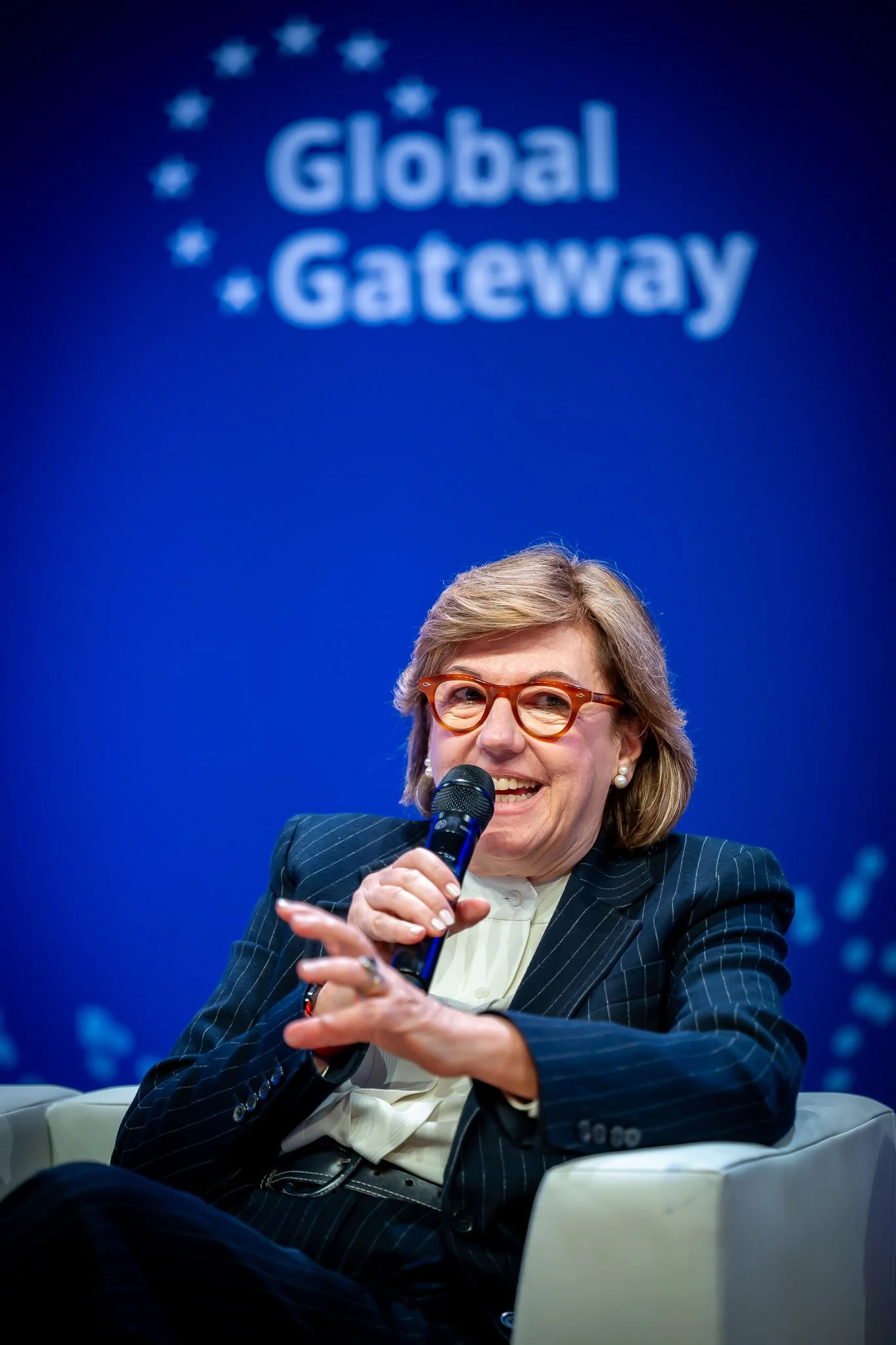 Une femme porte des lunettes et sourit en tenant un micro lors d'une conférence ou d'un événement sous un panneau avec le texte 'Global Gateway' en arrière-plan.