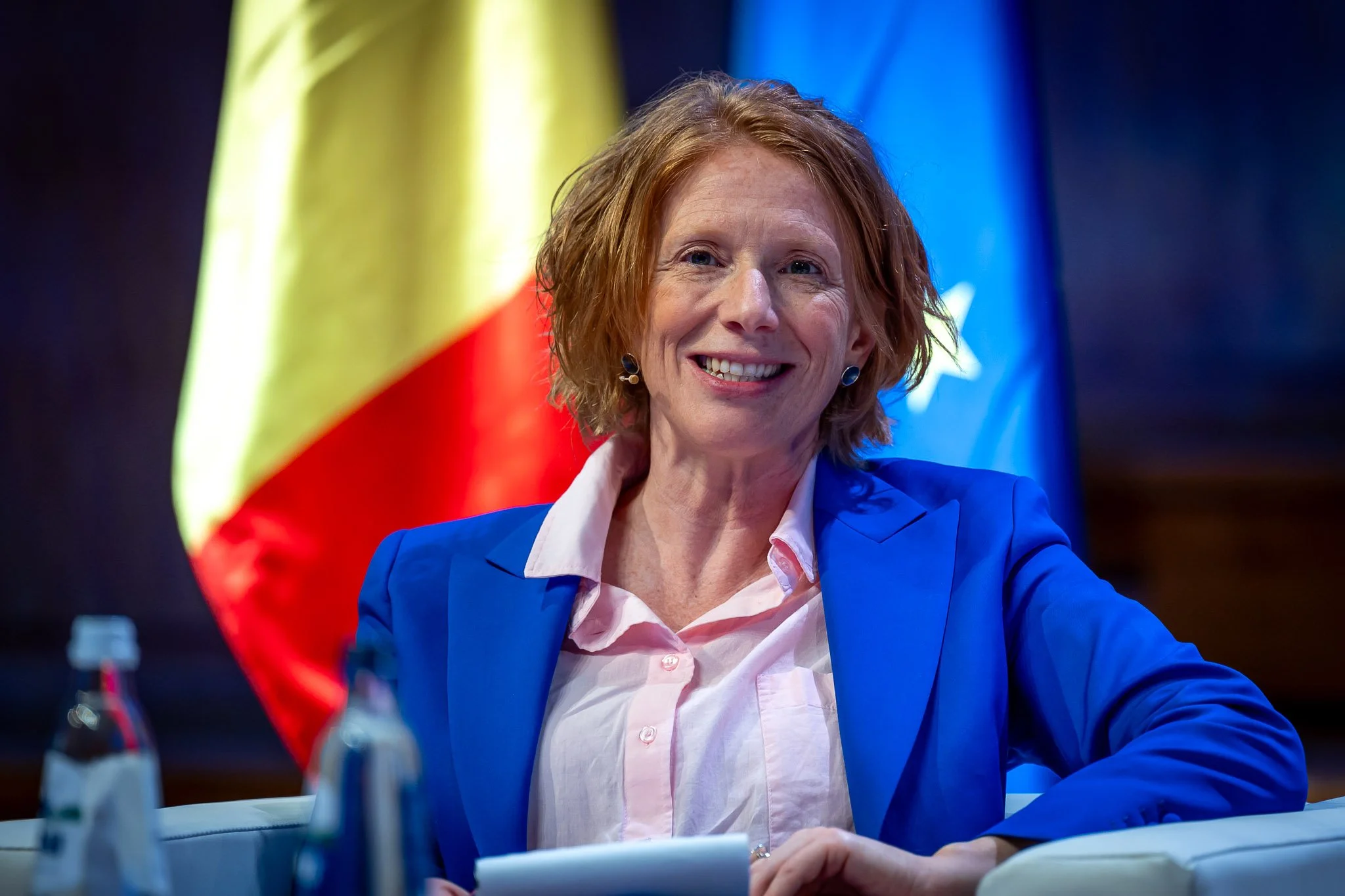 Une femme souriante avec des cheveux roux, portant une veste bleue et une chemise rose pâle, assise à une table avec des drapeaux en arrière-plan : un drapeau avec des couleurs jaune, rouge et vert, et un autre avec des étoiles. Elle semble être lors