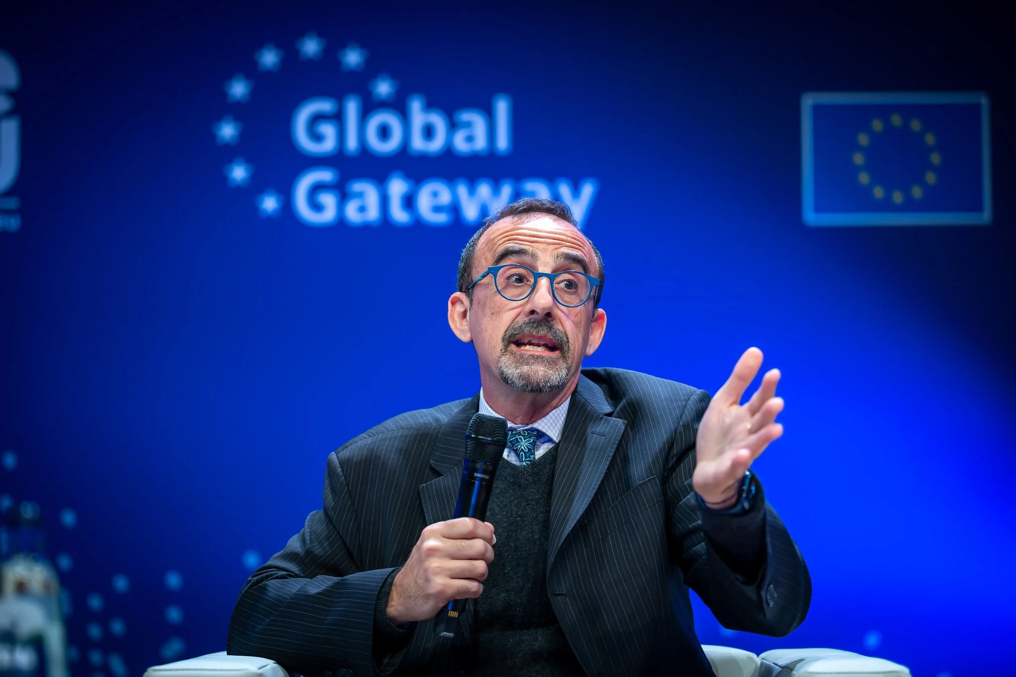A man in a suit holding a microphone at a conference, with the ‘Global Gateway’ logo and the European Union flag in the background.