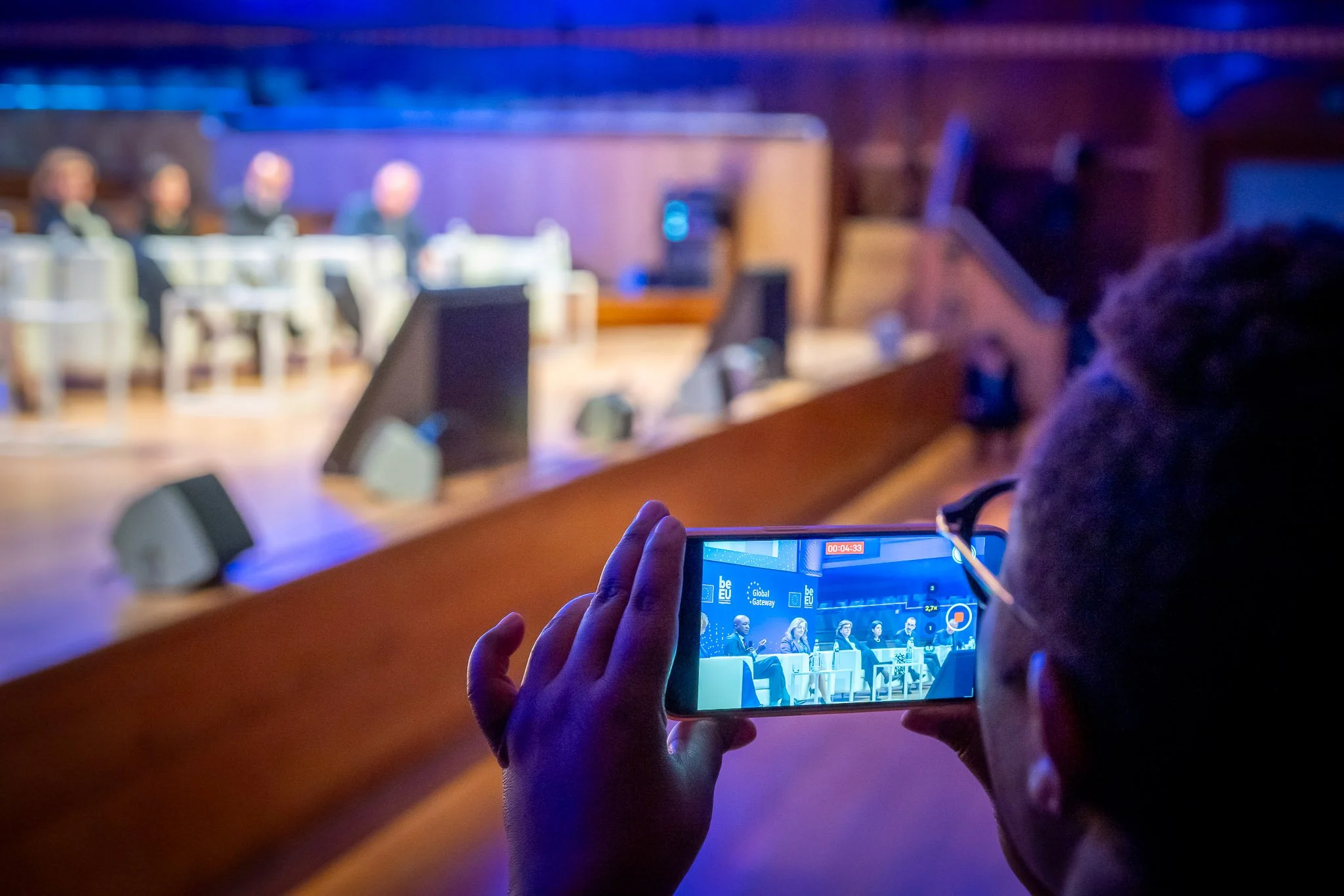 Une personne filme une conférence ou un panel à l'aide d'un smartphone en salle de réunion, avec plusieurs intervenants assis sur une scène en arrière-plan.