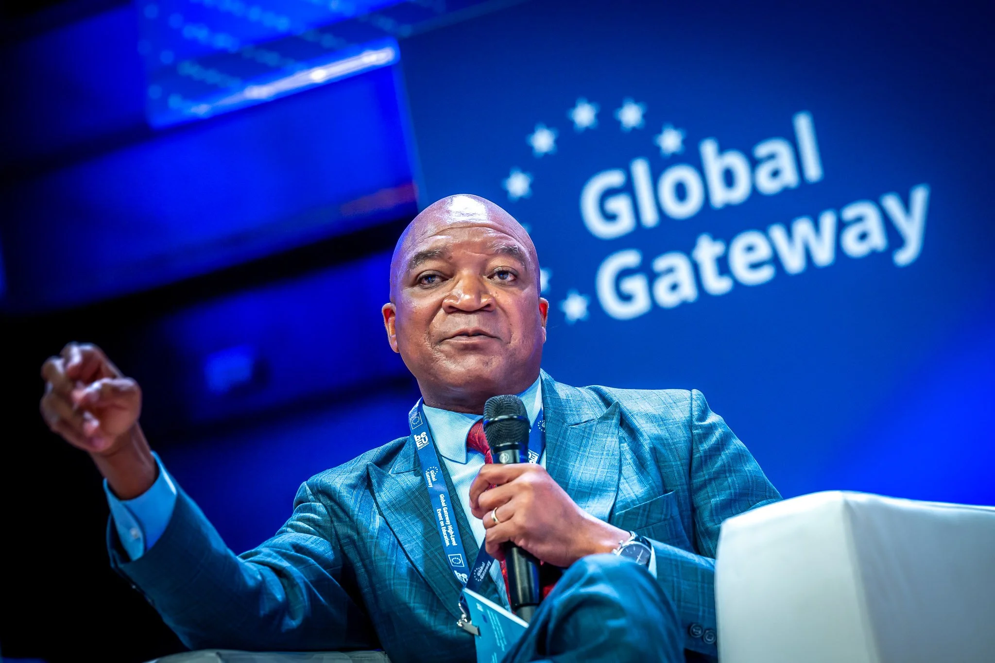 Beau portrait d'un conférencier en costume bleu parlant lors d'un événement "Global Gateway" avec un microphone en main, fond bleu avec le logo en arrière-plan.