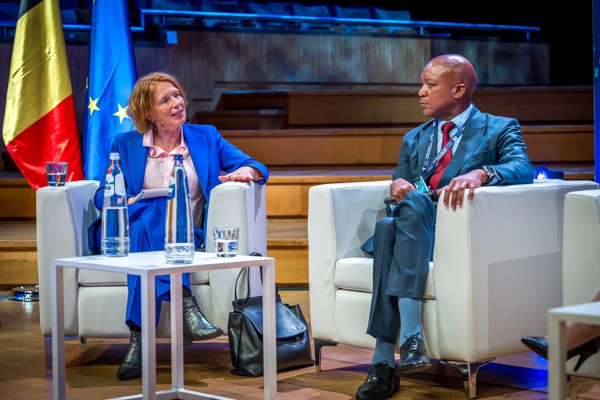 Two people sitting in armchairs at a conference, talking. Behind them are the flags of Belgium and the European Union. A table with bottles of water can be seen in the foreground.