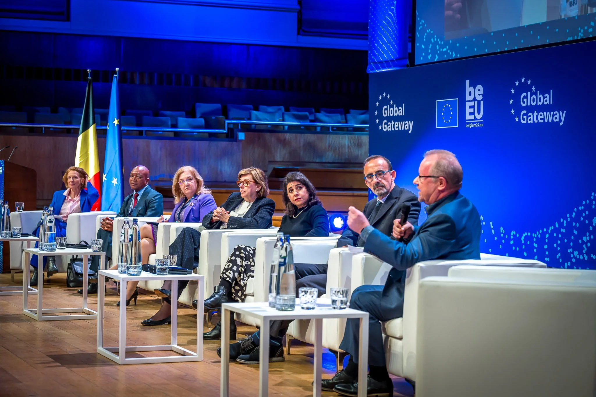 A conference scene showing several people seated on a stage, talking during a ‘Global Gateway’ event. European Union and Belgian flags in the background. The room is lit in blue.