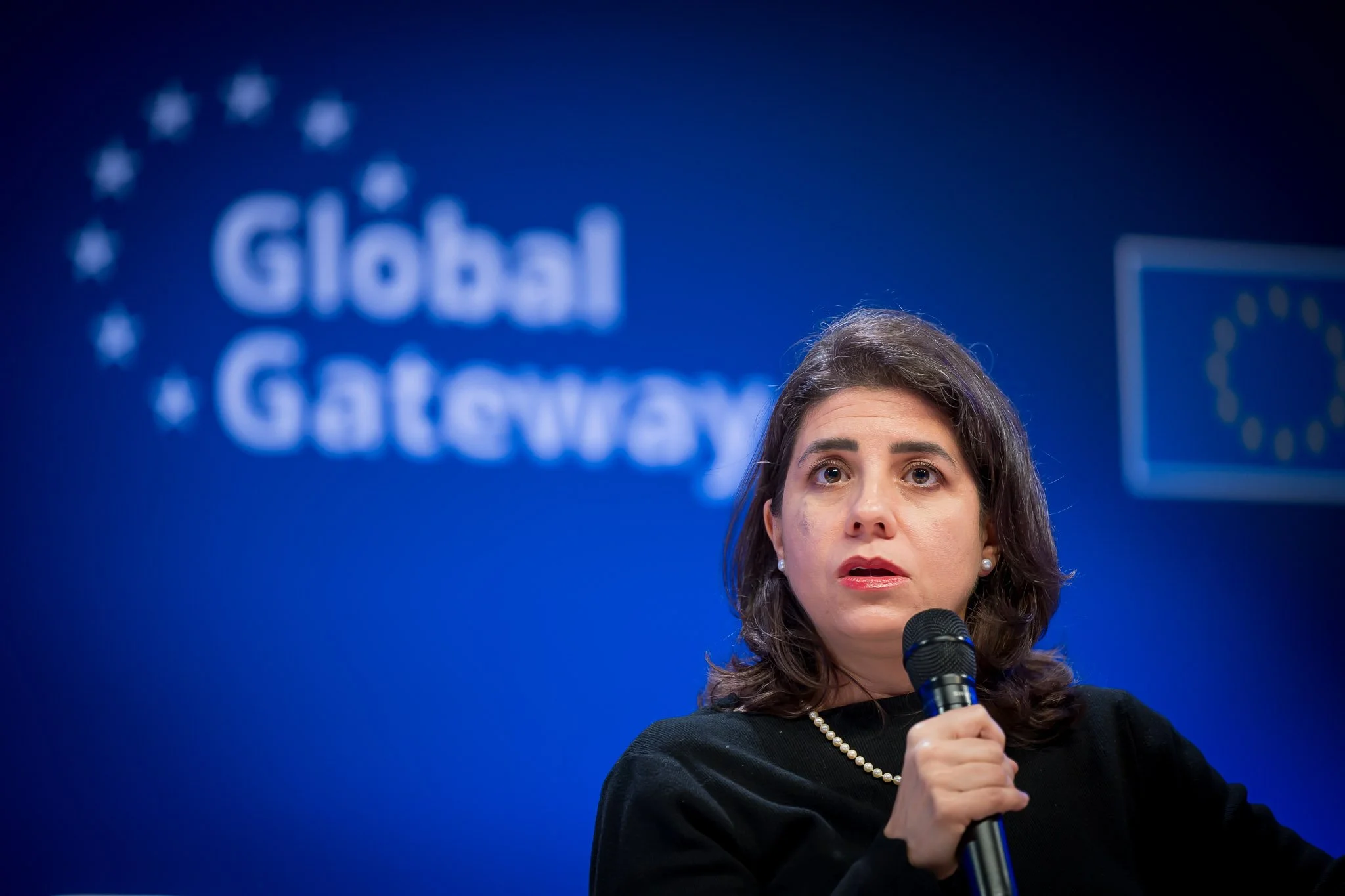 A woman holding a microphone, against a blue background featuring the text ‘Global Gateway’ and the European Union flag.
